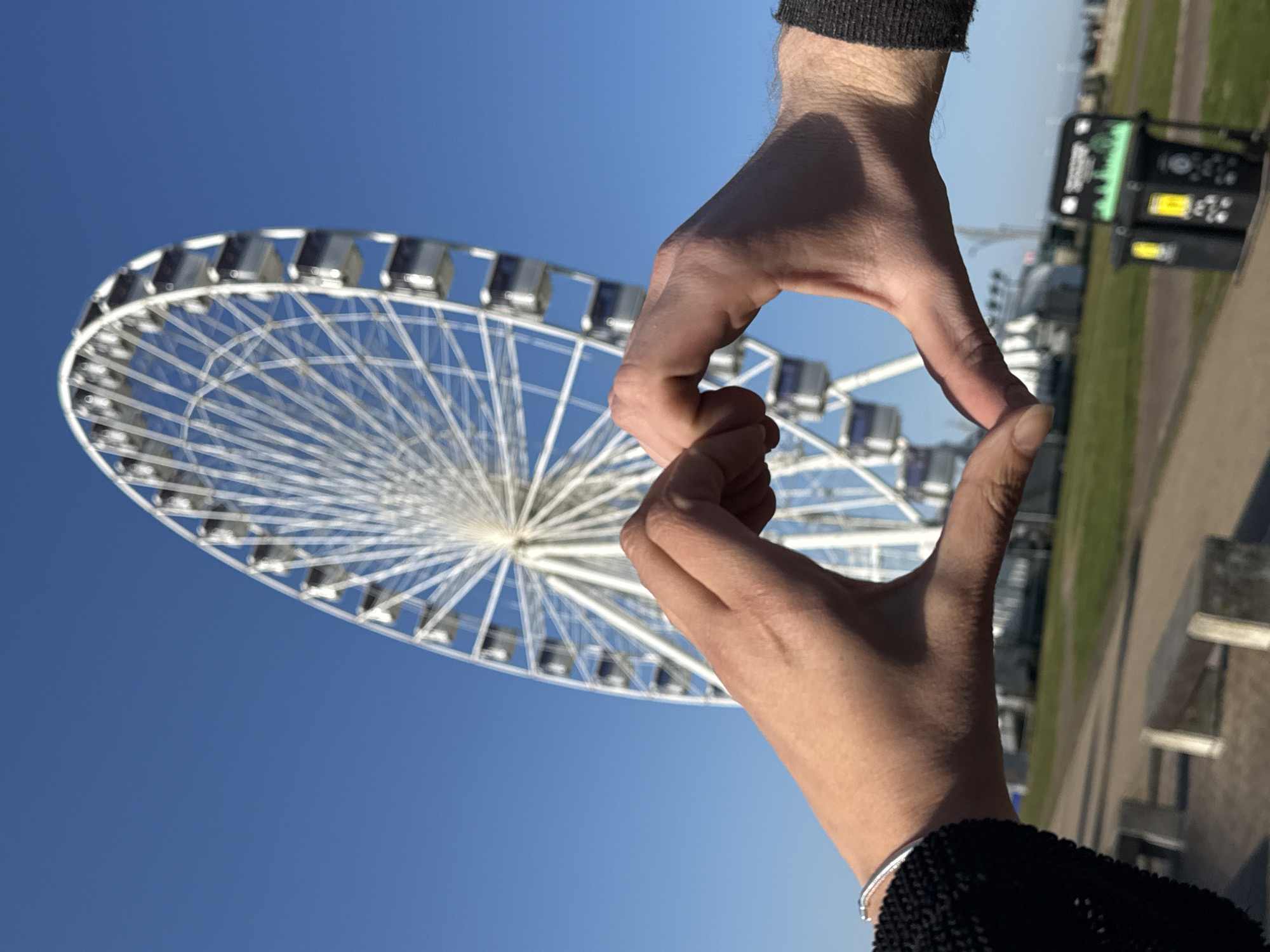 The image is a brightly lit, outdoor scene taken on a clear, sunny day, capturing a playful interaction with a prominent landmark. In the foreground, two hands are interlocked at the fingertips, forming a heart-like or diamond shape. The left hand, appearing more robust and possibly male, is clad in a dark sleeve, while the right hand, adorned with a silver bracelet and a dark knitted sleeve, is likely female. Their gesture symbolically frames the main subject in the background.
The central feature of the background is a large, white Ferris wheel, its intricate spokes and numerous enclosed gondolas clearly visible against a deep blue, cloudless sky. The wheel occupies a significant portion of the left side of the frame, suggesting it is a major attraction.
To the right of the hands, the scene includes a strip of green grass bordering a paved path or walkway. Further in the distance on the far right, a dark, rectangular public kiosk or machine stands, featuring a glowing screen that displays indistinct graphical elements, possibly sound waves or menu options, and various buttons. The vibrant blue sky dominates the upper and central parts of the image, indicating excellent weather conditions. The bright sunlight casts distinct shadows, confirming it is daytime, likely mid-day. The overall setting points to a popular tourist or leisure area, consistent with Great Yarmouth, United Kingdom. No specific text is clearly legible on the kiosk.