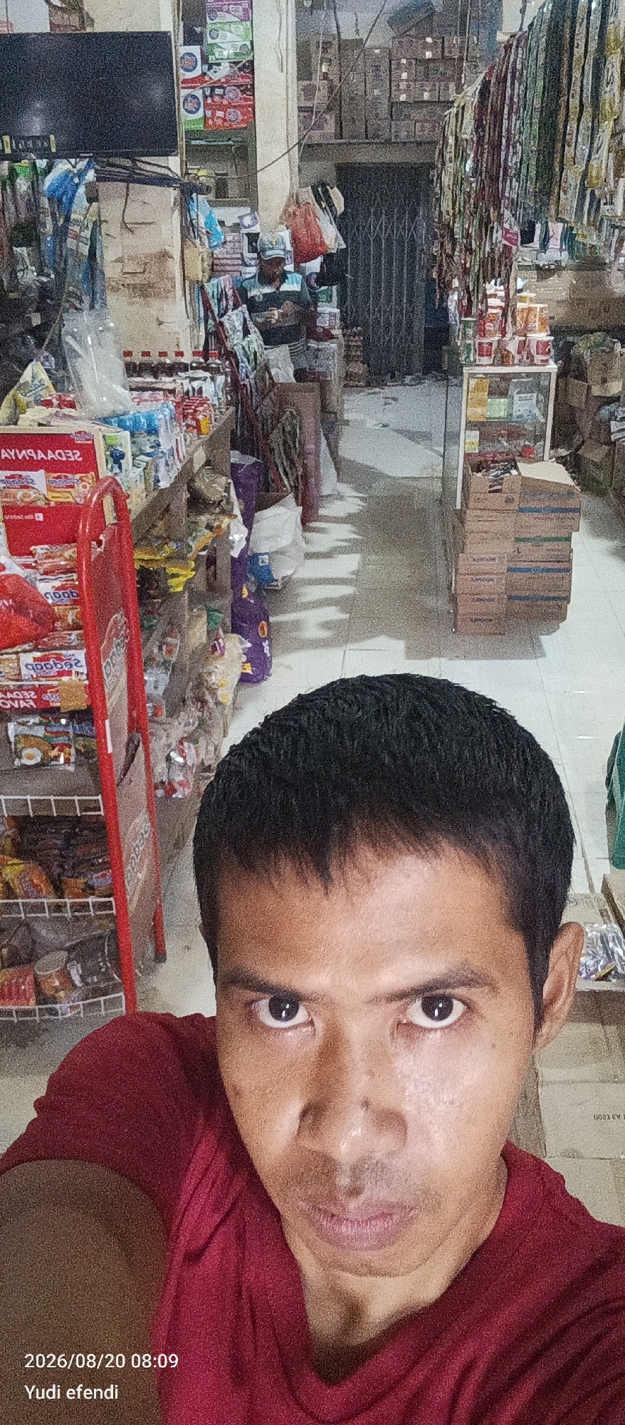 A close-up, selfie-style shot shows a man's face filling the foreground. He has short, dark hair and is wearing a red shirt. His eyes are open and looking directly at the camera. In the background, a convenience store is visible. Shelves are stocked with various packaged goods, including snacks and drinks. A red display rack stands to the left, and further back, a metal gate is closed. The store appears to be located in Rawajitu Selatan, Indonesia, as indicated by the timestamp and name "Yudi efendi" in the bottom left corner of the image, which is a common format for date and time stamps on photos taken in Indonesia. The overall scene suggests an informal retail environment.
