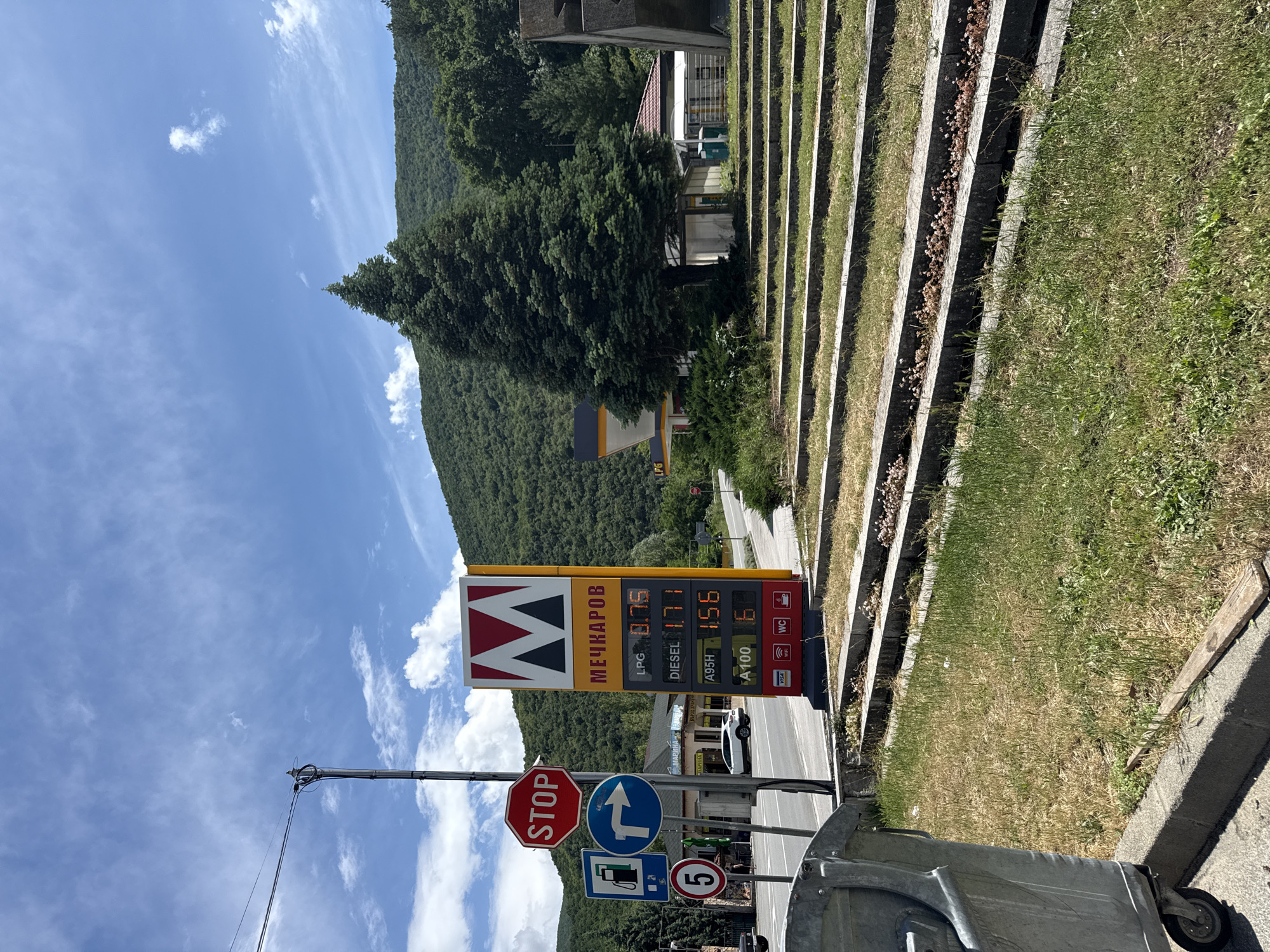 The image, taken in Vetren Dol, Bulgaria, depicts a sunny daytime scene featuring a roadside gas station against a mountainous backdrop. In the foreground, a gray wheeled trash can is visible on the right. A utility pole with power lines extends from the left.
Dominating the midground is a tall yellow and red gas station sign, displaying the name "МЕЧКАРОВ" (MECHKAROV) in Cyrillic and a distinctive "M" logo. Current fuel prices are listed: LPG at 0.75, DIESEL at 1.71, A95H at 1.56, and A100 at 6. The sign also indicates available services with icons for WC, WiFi, and acceptance of VISA cards. Behind the sign, a white car is partially visible.
To the left of the gas station sign, several road signs are mounted on a pole: a red octagonal "STOP" sign, a blue circular sign indicating a right turn, a blue rectangular sign showing a gas pump, and a blue circular sign with the number "5".
On the right side of the image, a series of concrete steps with grass growing between them ascends a slope. Further back, buildings with red roofs are nestled among trees. The background features lush green mountains under a bright blue sky dotted with scattered white clouds, suggesting clear and pleasant weather. The overall scene is one of a functional roadside stop integrated into a natural, scenic environment.