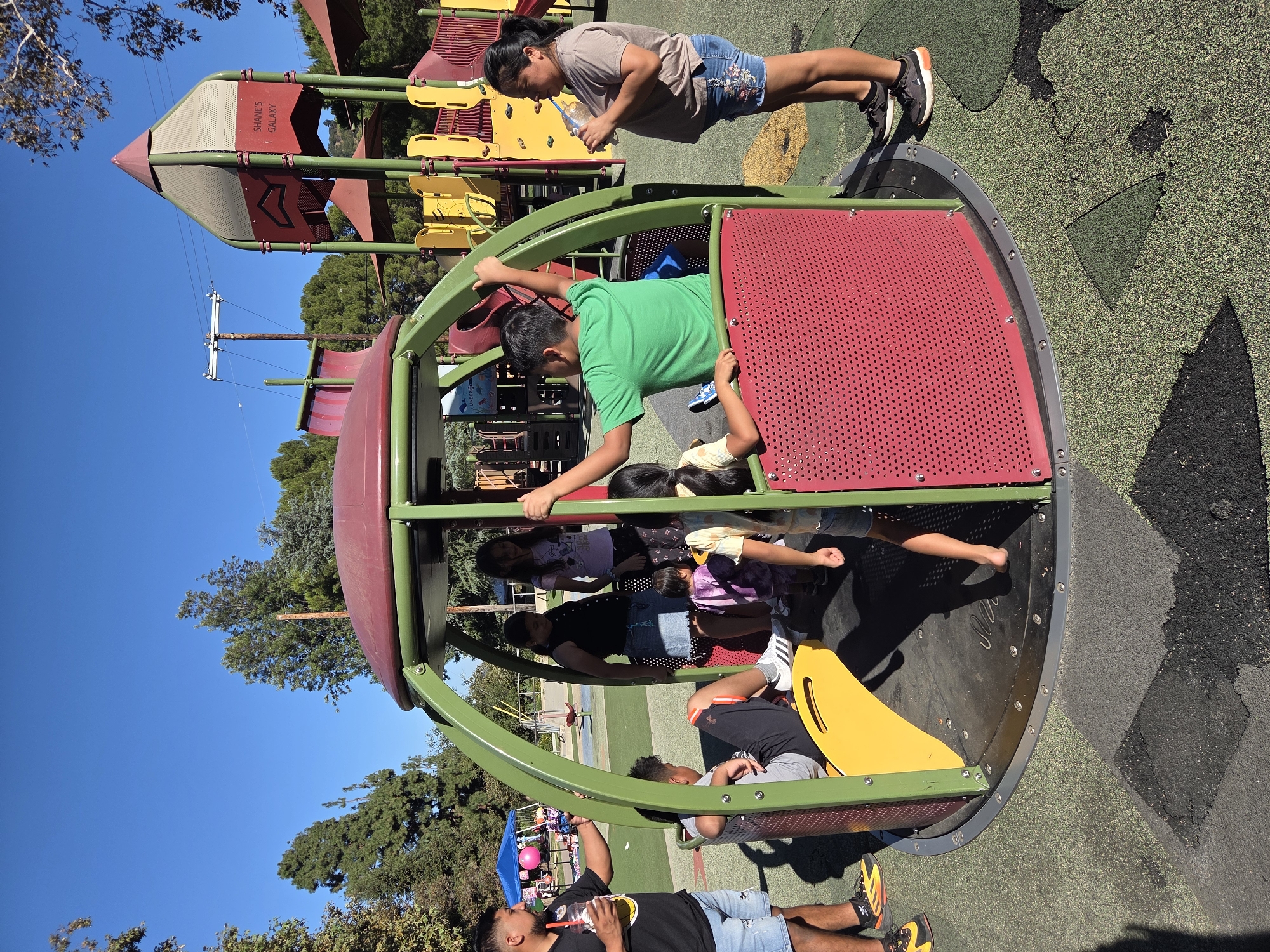 This is a medium shot, taken outdoors, of a diverse group of children and two adults on a playground on a sunny day. The playground is set within a park with lush green trees and a clear blue sky. The central focus is a multi-person merry-go-round with green metal framing and red perforated panels. Inside and around the merry-go-round, six children are visible. One boy in a green t-shirt is in the center of the structure, appearing to be operating or interacting with it. Two girls, one in a dark top and denim skirt, and another in a light purple t-shirt, are standing. A younger child in a purple outfit is near them. Another child's back is visible, wearing a light-colored shirt. An adult male, wearing a black t-shirt and denim shorts, stands to the left of the merry-go-round, holding a cup with a yellow beverage. To the right, an adult female, in a beige t-shirt and floral denim shorts, drinks from a water bottle. In the background, there's another playground structure with red and yellow elements, a blue tent, and some trees. The ground is a resilient, multi-colored rubberized surface. The overall scene depicts a playful and social atmosphere.