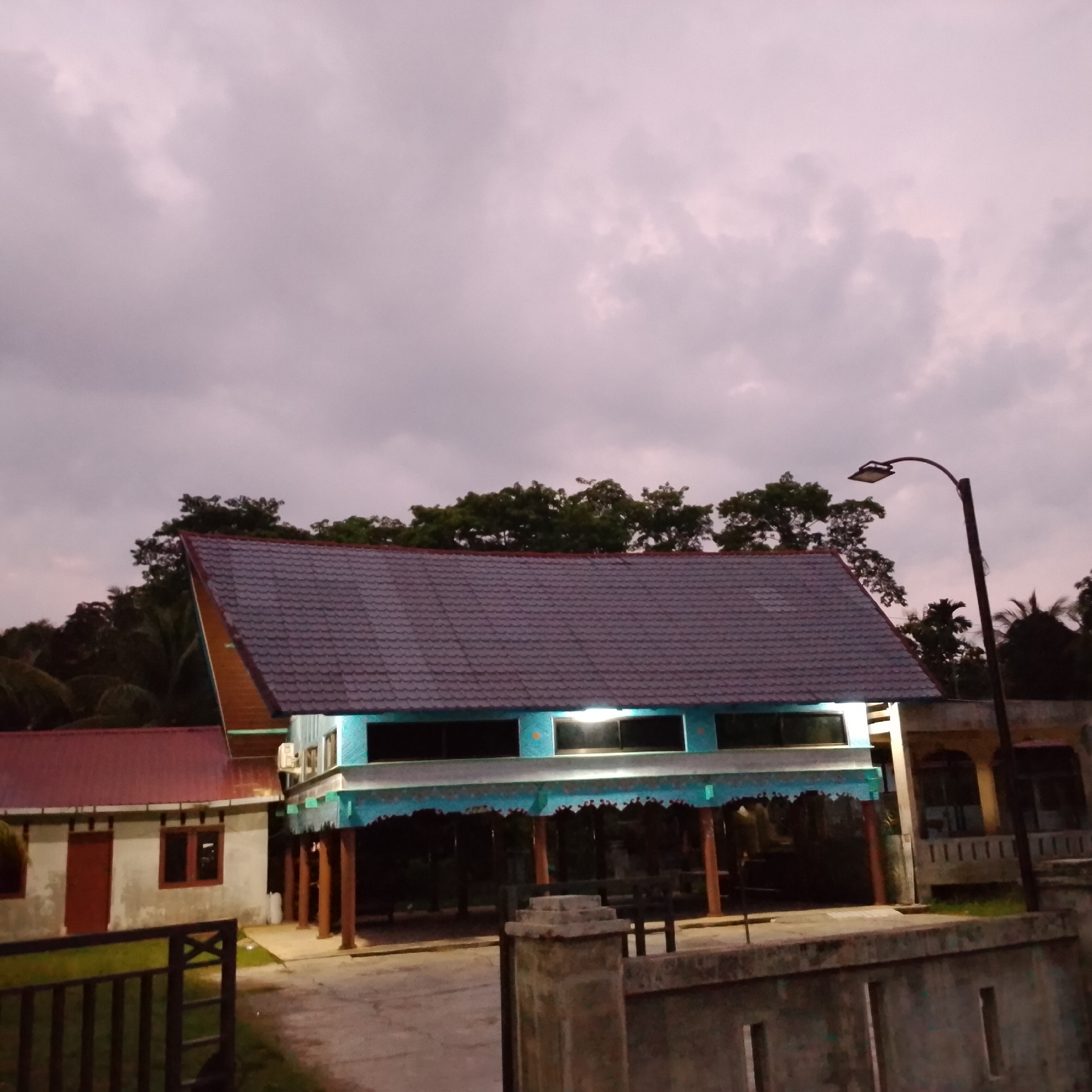 The image captures an outdoor scene at dusk, featuring a traditional house with a distinctive blue and teal elevated structure supported by wooden stilts. This structure has a dark-tiled, angled roof and features large windows with interior lighting. Adjacent to it, a smaller building with a red roof has two windows and a red door. A dark metal fence is visible in the foreground to the left. A street lamp stands to the right of the elevated house. The background is filled with dense green trees, and the sky above is overcast with a dusky hue, suggesting either early morning or late evening. The scene appears to be in a rural or semi-rural area, given the presence of trees and the architectural style of the main house.