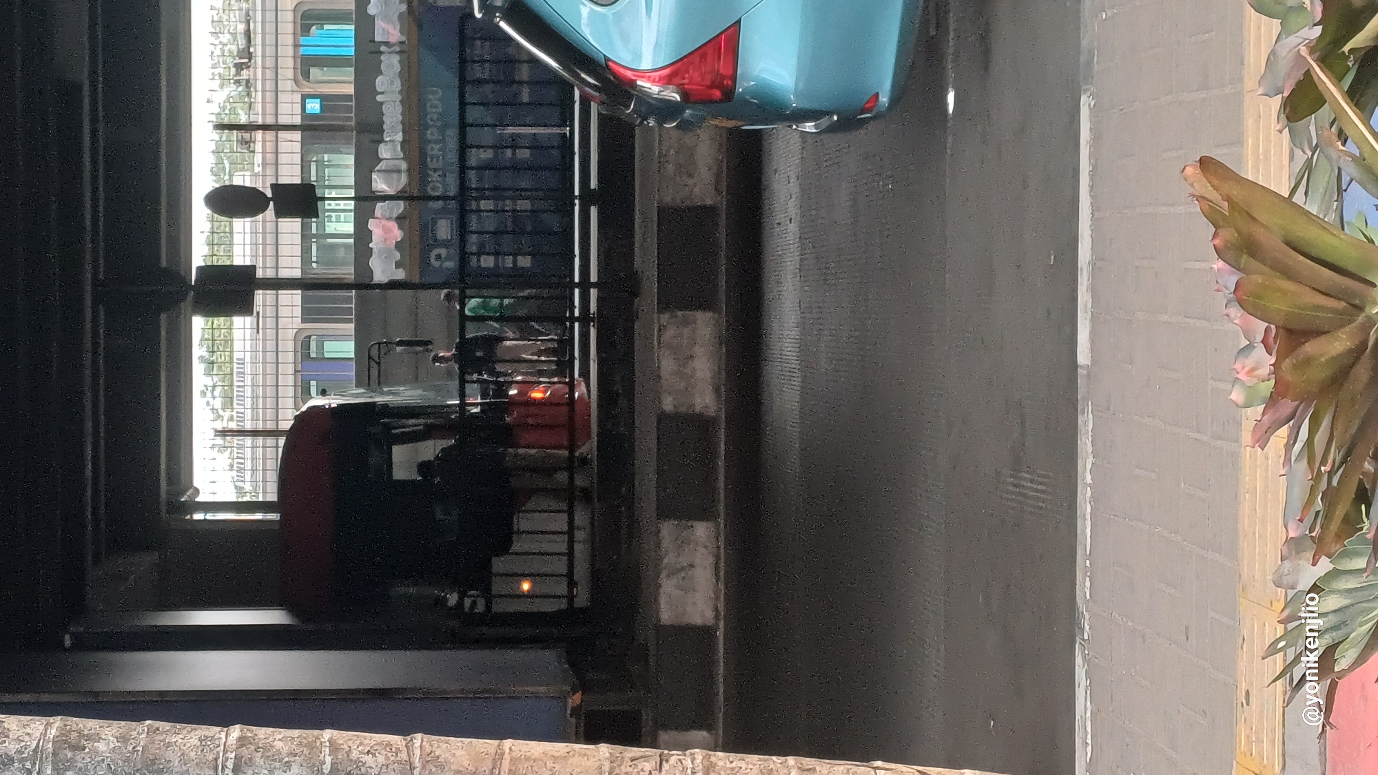 The image depicts an outdoor scene in South Jakarta, Indonesia, likely near a transportation hub. In the foreground, a light blue car is partially visible, facing away from the camera. Behind the car, a road surface with visible speed bumps and a sidewalk can be observed. Further back, a black metal fence runs horizontally, separating the road area from an elevated structure. Through the fence, a red bus and several individuals, appearing to be passengers or workers, are visible. Behind the bus, a train with blue and white carriages is stopped at a platform, indicated by the presence of tracks and station architecture. The overall environment suggests a busy urban area with multiple modes of public transportation. The lighting indicates it is daytime.