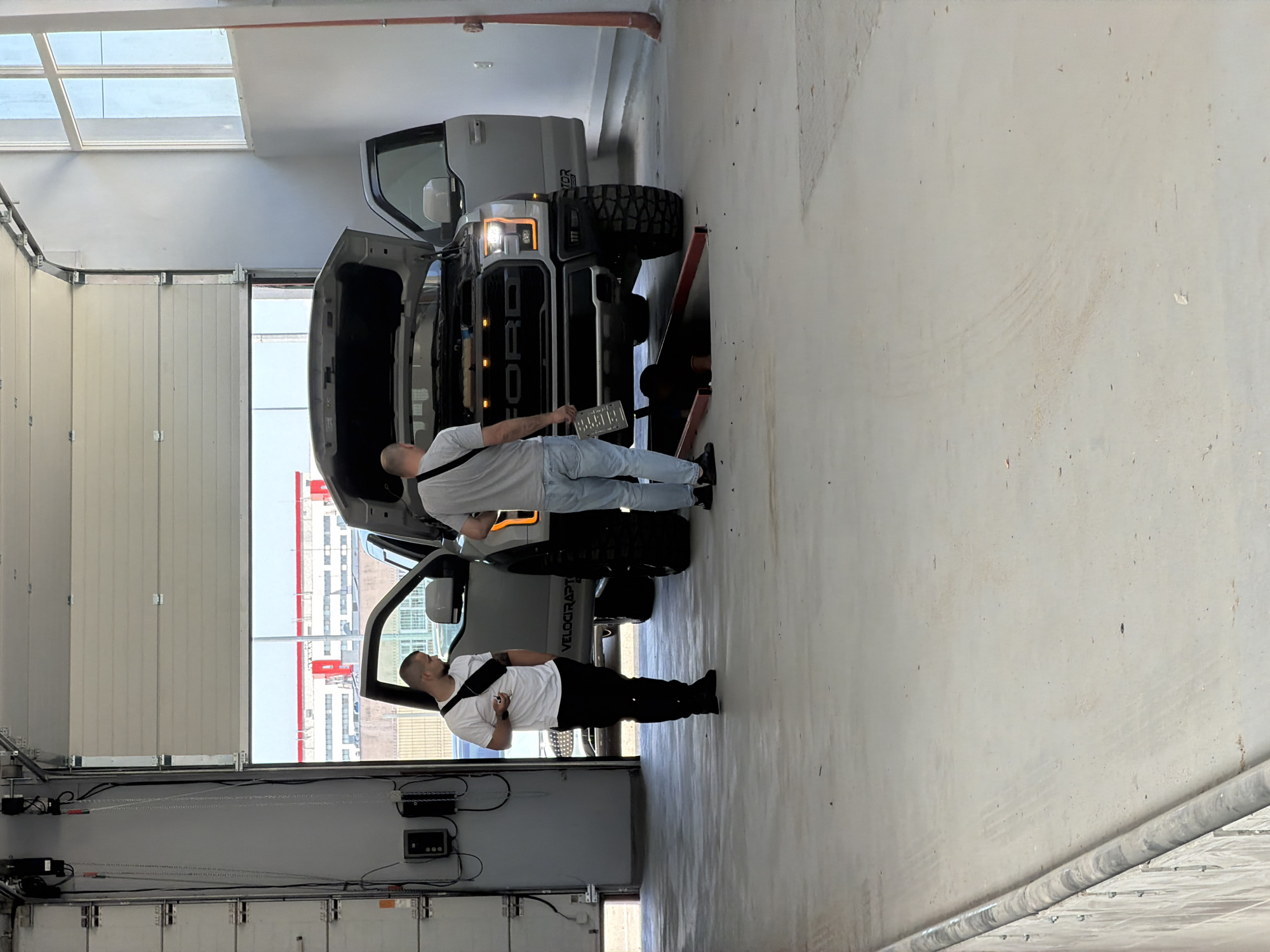 Two men are standing in what appears to be a workshop or garage with a large overhead door open to the outside. In the center of the scene is a Ford F-150 Raptor pickup truck with its hood open. One man, wearing a white t-shirt, black pants, and a black fanny pack, stands to the left of the truck, holding a cigarette. The second man, wearing a gray t-shirt and blue jeans, stands near the front of the truck, holding a license plate. The truck has large, aggressive off-road tires. The floor is concrete and shows some tire marks. The background outside the open door reveals industrial buildings and a clear sky. This scene is located in the city of Ruse, Bulgaria.