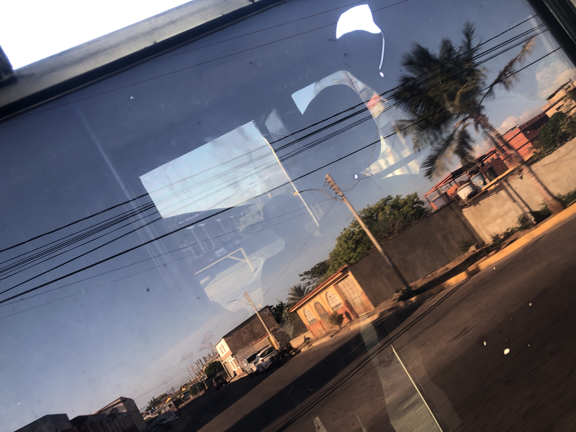 The image, likely taken from inside a moving vehicle, shows a partial view of Juan Griego, Venezuela, through a window. The window reflects the interior of the vehicle. Visible on the right is a palm tree. The cityscape includes buildings of varied architectural styles, including a beige building with arched windows. Several vehicles are parked along the street. Power lines run across the sky. The overall scene conveys a sense of everyday life in a tropical urban environment.