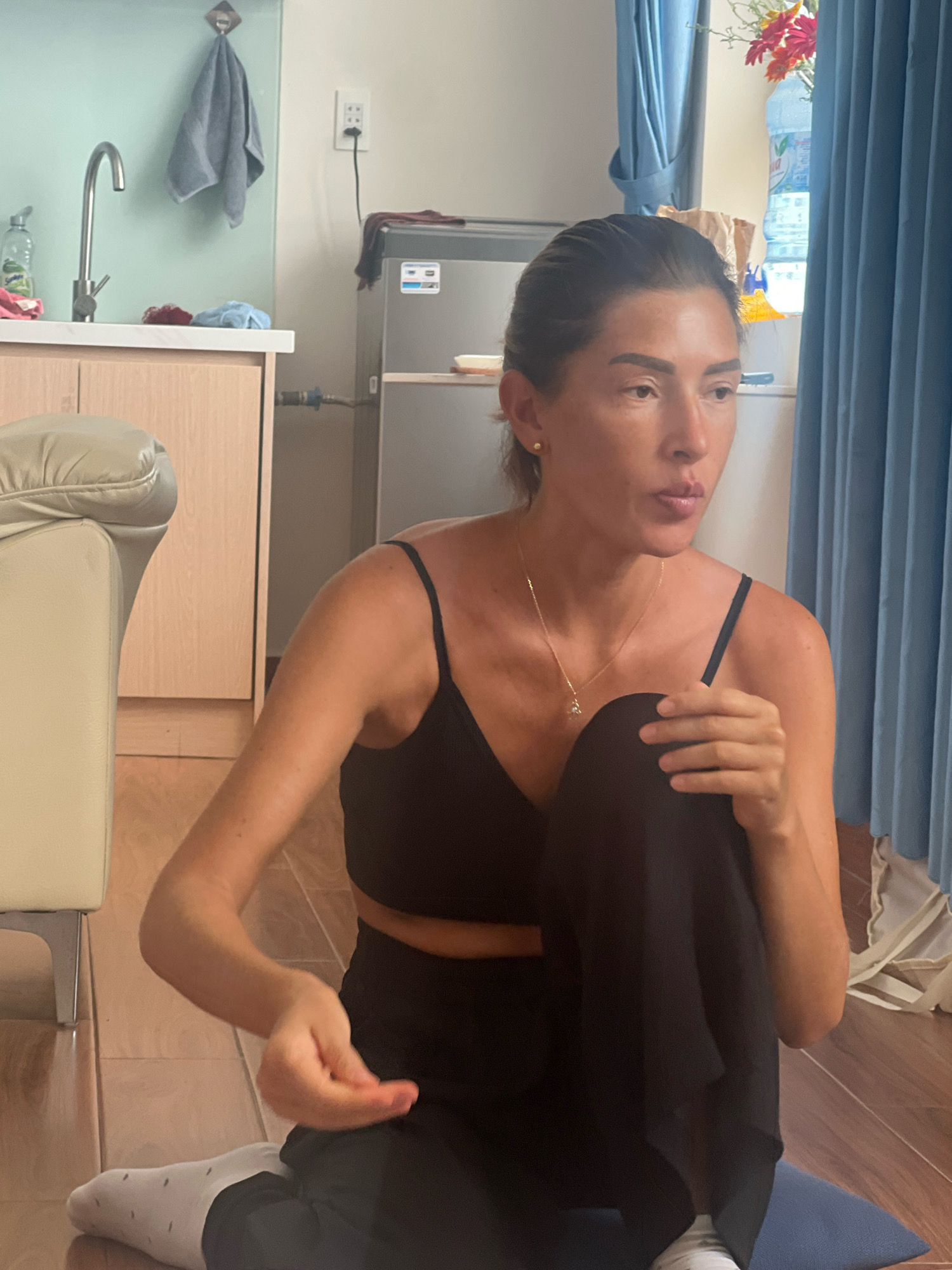 The image shows a woman sitting on the floor in what appears to be an apartment in Đà Nẵng, Vietnam. She is wearing a black sports bra and black pants, with white socks. Her hair is pulled back, and she is wearing a gold necklace with a pendant. The room has tiled flooring and a wooden cabinet with a sink and faucet on the left. In the background, there is a refrigerator and blue curtains. The woman is looking to the right, and her hands are resting on her lap, holding a piece of black fabric.