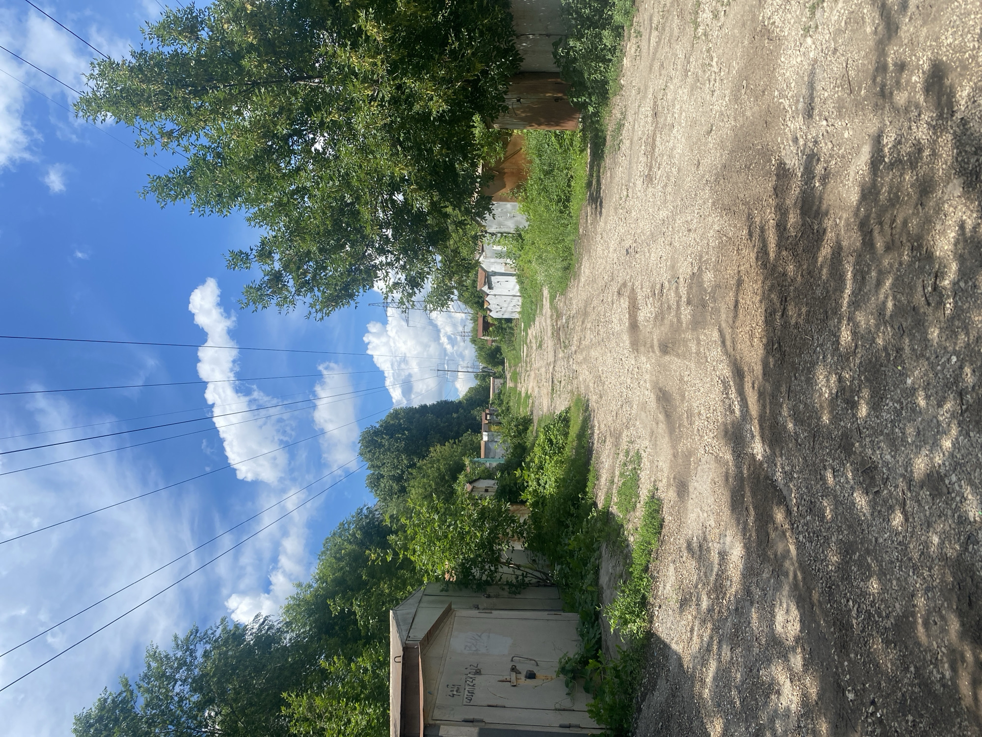 The image depicts a sun-drenched, unpaved road in a rural or suburban area, likely in Samara, Russia. The road is lined with trees and overgrown vegetation, with several small, weathered garages or sheds visible on the left side. The garages appear to be made of metal and concrete, and some have visible graffiti. Overhead, power lines crisscross the bright blue sky, which is dotted with fluffy white clouds. The lighting suggests it is daytime, possibly late morning or early afternoon. The overall impression is one of a quiet, somewhat neglected but lived-in setting on a warm, clear day. There is no discernible activity or people in the image. Some faint text is visible on one of the garages, appearing to be a serial number or code.
