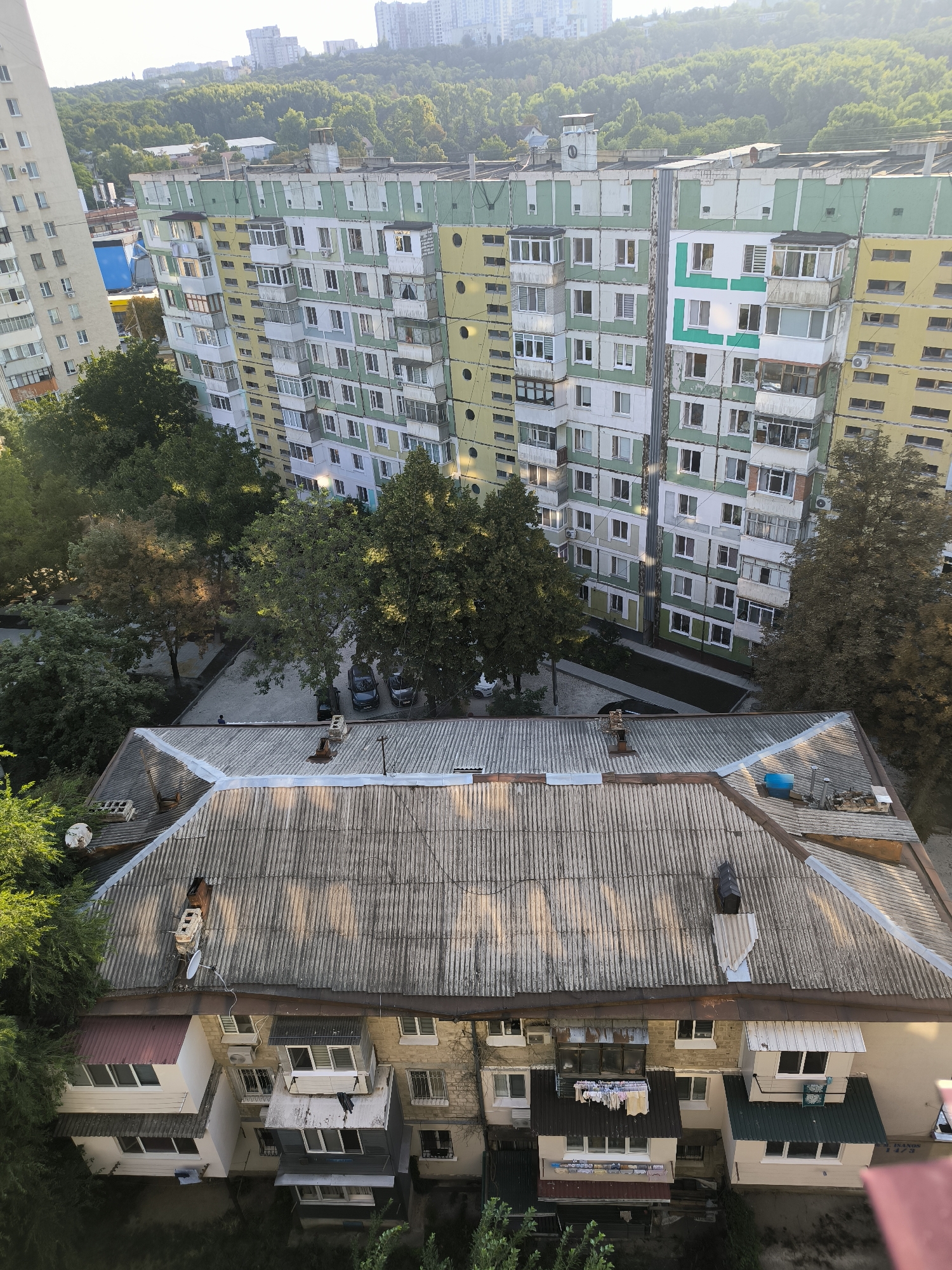 The image, taken from a high vantage point, showcases a view of residential buildings in Chișinău, Moldova, bathed in the warm light of what appears to be late afternoon or early evening. In the foreground, the weathered, corrugated metal roof of a lower building dominates, showing signs of age and wear. Several chimneys and ventilation pipes protrude from this roof, along with a satellite dish. The building beneath this roof features multiple stories with a mix of window styles, some with air conditioning units, and small balconies. Laundry is visible hanging on a line on one of the balconies.
Behind this foreground structure, a larger, more imposing apartment block stands, painted in alternating bands of green, yellow, and white. This building exhibits numerous windows, many with enclosed or semi-enclosed balconies, characteristic of Soviet-era architecture common in Chișinău. Some windows are illuminated, suggesting occupancy. A striking feature of this building is a large, stylized green "L" shape painted on a white section of its facade. Adjacent to this building, another similar apartment block, predominantly yellow, is partially visible.
The area around these buildings is dotted with mature trees, their foliage creating a dappled effect on the ground below, where a few parked cars are visible in what appears to be a courtyard or parking area. In the far distance, a hazy skyline of taller, more modern buildings is discernible against a bright, possibly sunlit, sky, suggesting a broader urban landscape beyond this immediate neighborhood. The overall impression is of a densely populated urban environment with a mix of older and newer architectural styles, set against a backdrop of greenery. The presence of what appears to be a lone individual standing on the roof in the middle ground, between the two main building types, adds a human element to the scene.