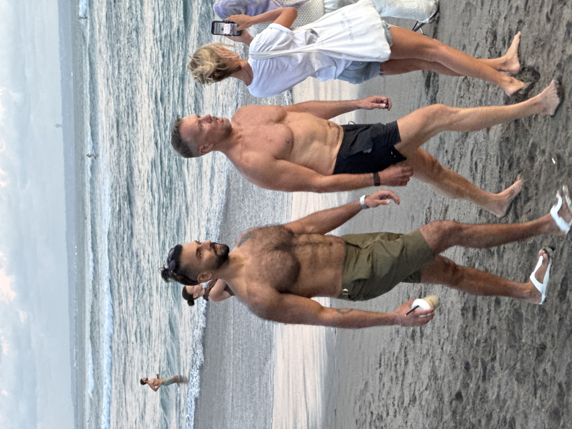 The image shows three adults on a beach in North Kuta, Indonesia. Two men, one wearing black shorts and the other wearing khaki shorts, are walking in the foreground. The man in khaki shorts is holding a beverage. Behind them, a woman in a white dress is taking a photo with her phone. In the background, a person is swimming in the ocean. The scene includes the beach, ocean, and a cloudy sky.