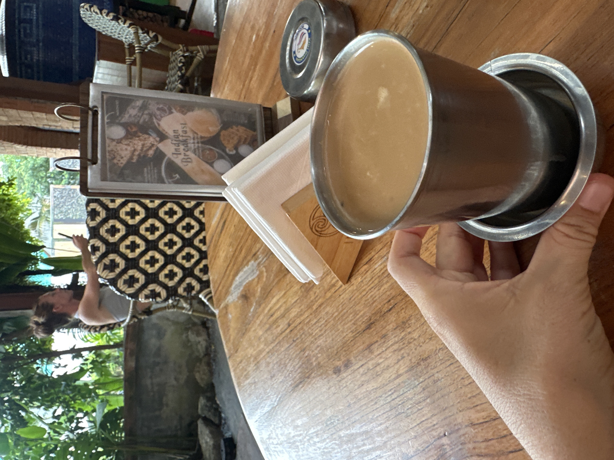 A hand holds a metal cup of coffee on a wooden table in an outdoor cafe. In the background, a woman sits on a chair and looks at her phone. A menu board is also visible on the table. The cafe appears to be located in a tropical environment with lush greenery.