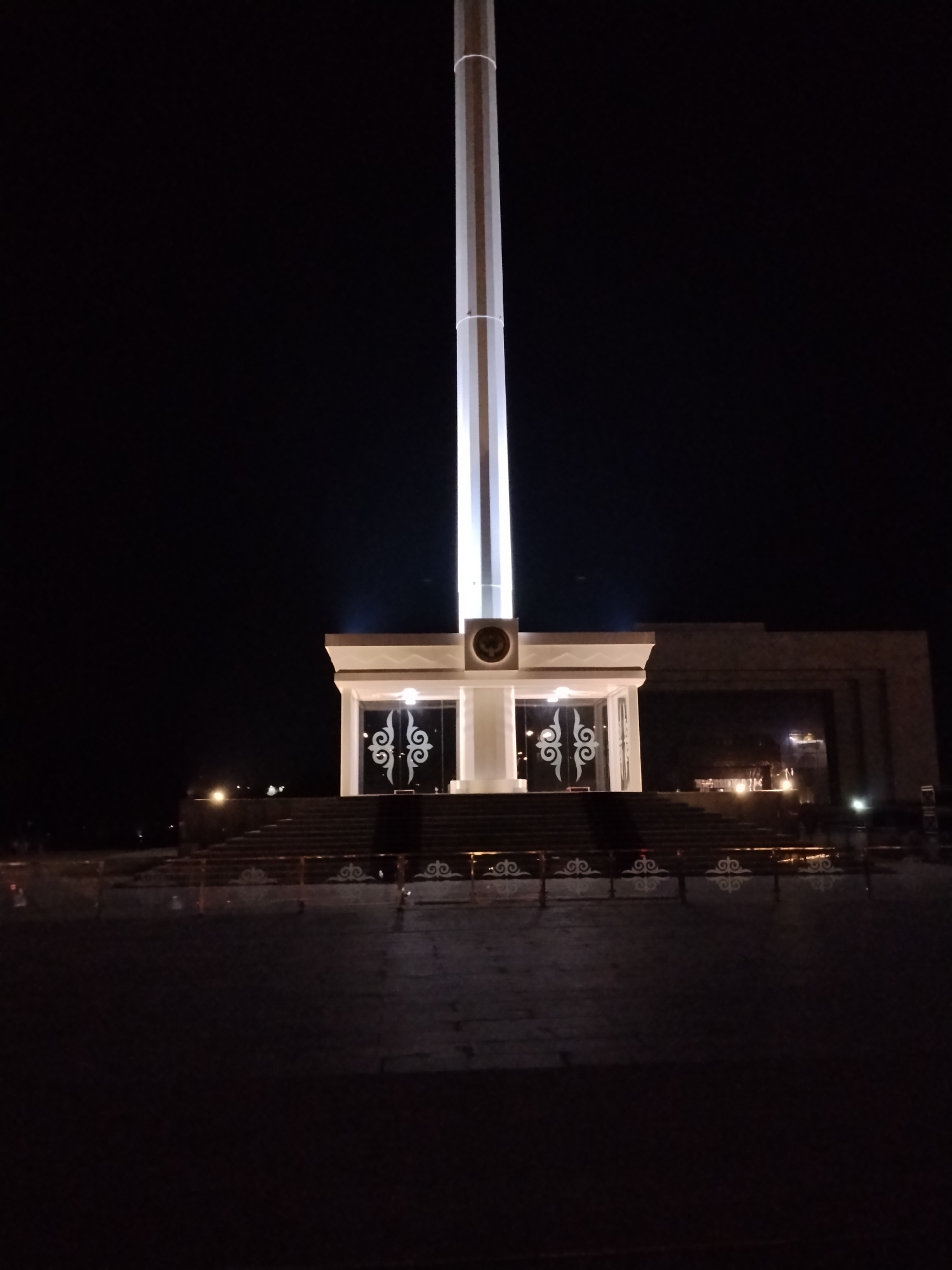 The image shows the Monument of Freedom in Bishkek, Kyrgyzstan, at night. The monument consists of a tall, white flagpole illuminated by lights. At the base of the flagpole is a modern structure with glass doors featuring decorative patterns and a circular emblem above the doors. A wide set of stairs leads up to this structure. The surrounding area appears to be a paved plaza. The sky is dark, indicating nighttime. No people are visible in the frame.