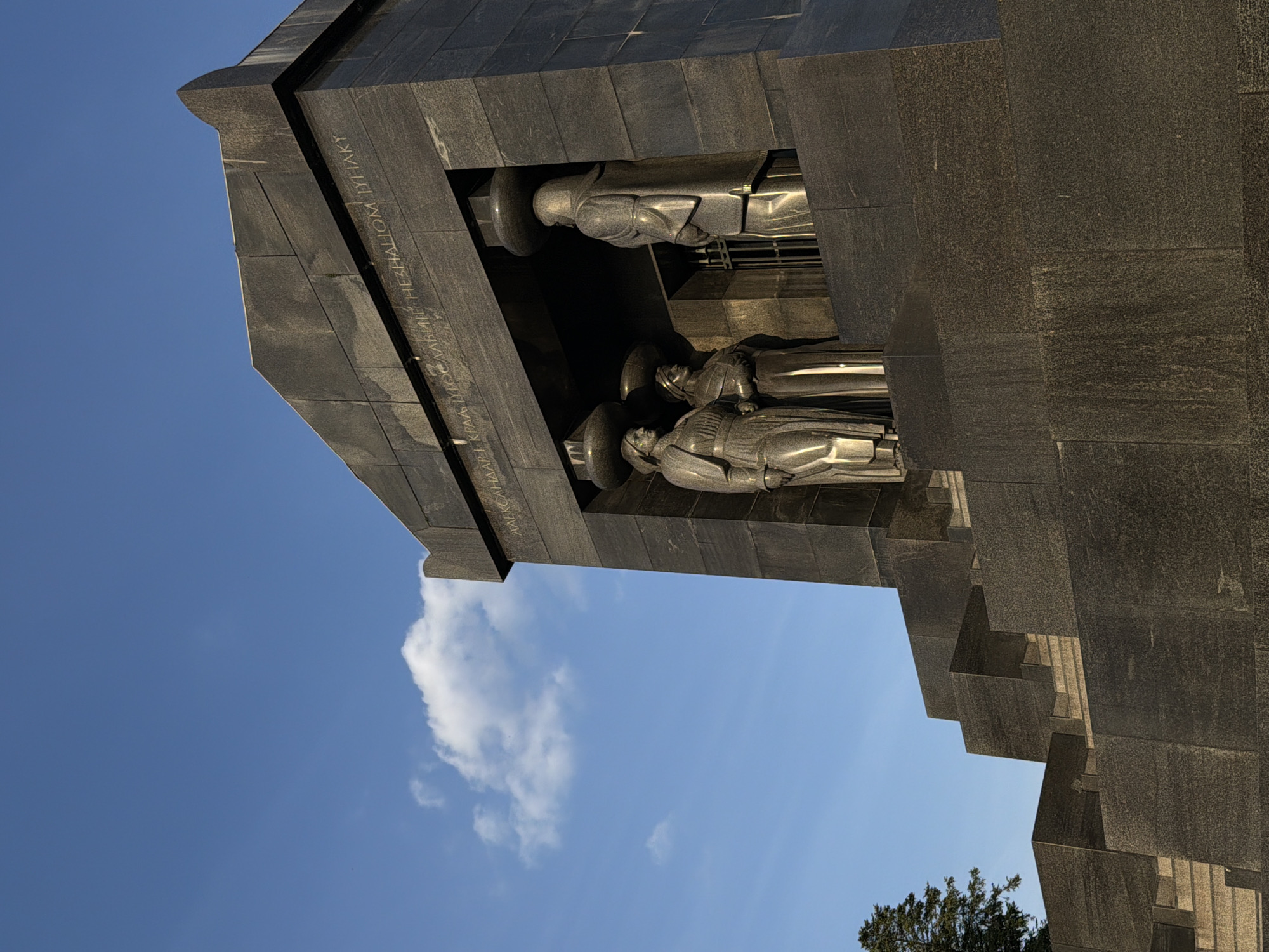 The image captures a portion of the Monument to the Unknown Hero on Avala Mountain in the Vozdovac Urban Municipality, Serbia. Visible are sections of the monument's dark, stone architecture against a backdrop of a blue sky with white clouds. Three sculptural figures are positioned within a recessed section of the monument. Their orientation suggests they may be representations of fallen soldiers or heroes. The monument's facade is engraved with Cyrillic lettering. The overall impression is one of a solemn and historical landmark.