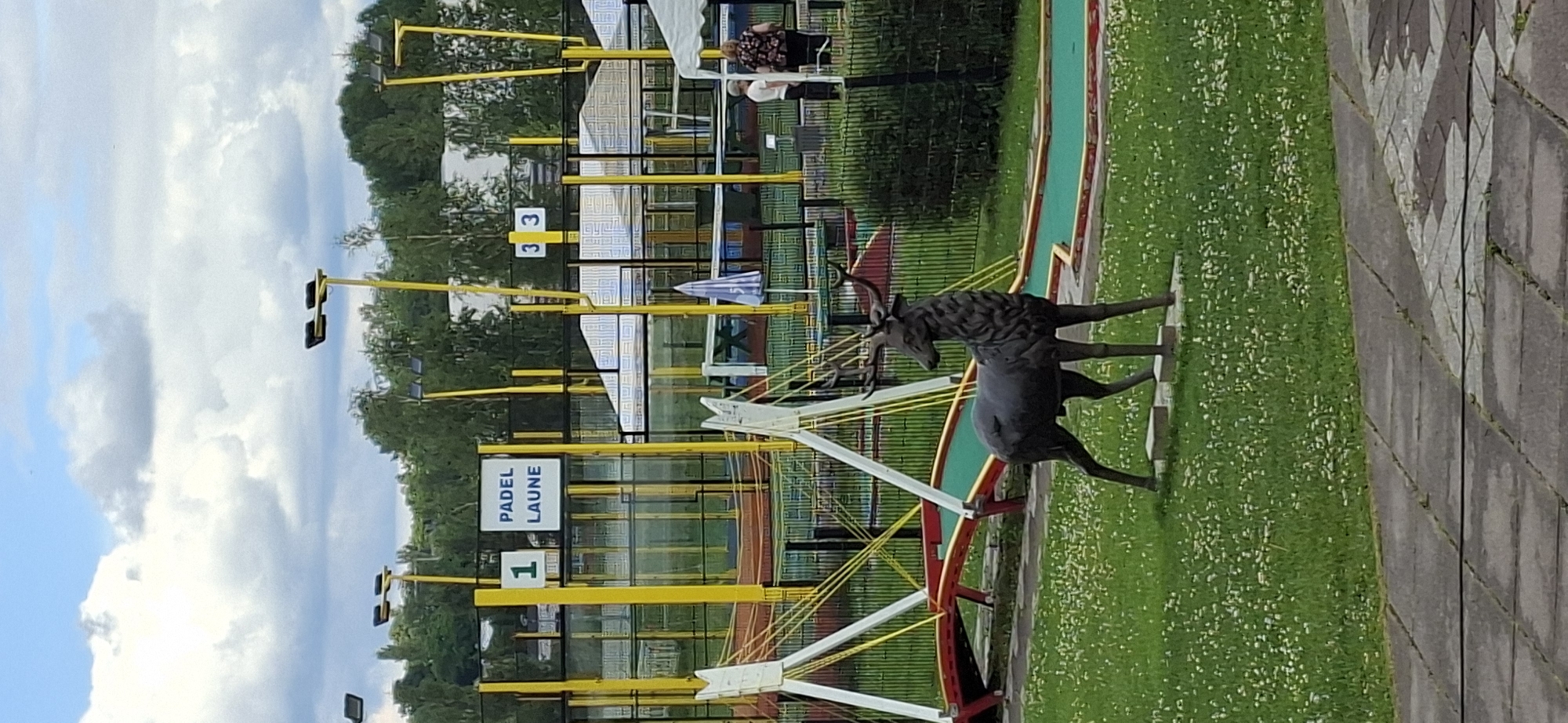 A statue of a deer stands on a grassy area dotted with small white flowers. Behind the deer, a mini-golf course is visible with green turf and red and yellow borders. Several yellow poles with lights stand around the course. A sign in the background reads "PADEL LAUNE" and has the number "1" to its left. Further to the right, another sign shows the numbers "3 3". In the far background, trees form a dense line. Two people are visible near a fence in the middle ground, one in a white shirt and the other in a floral patterned top. A blue umbrella is also present on the mini-golf course. The sky is partly cloudy. The location is Padel Laune in Lahti, Finland.
