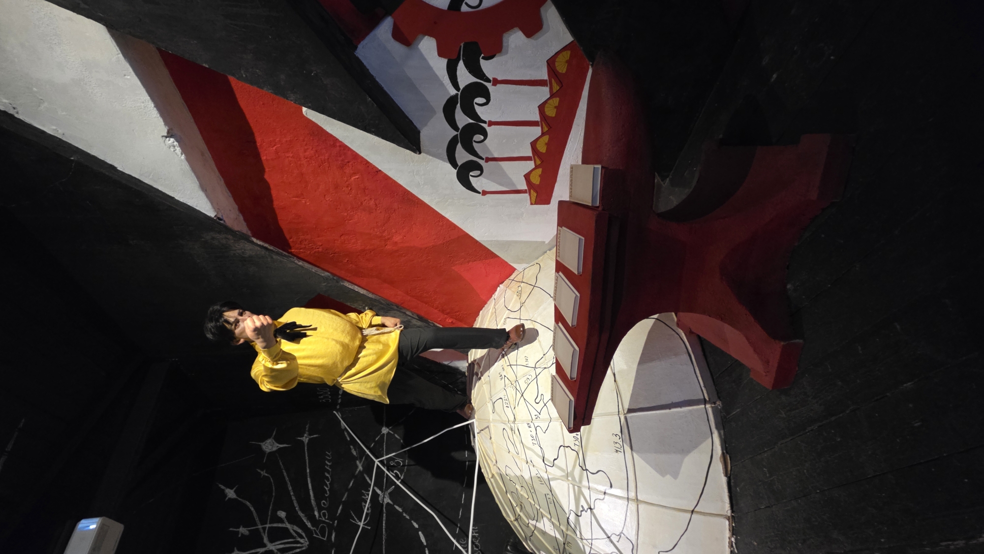 The image shows a man standing in an unusual space in Saint Petersburg, Russia. He is wearing a yellow shirt and black pants and is holding a microphone in his left hand. The man appears to be giving a presentation or performance.
The background consists of a complex architectural design, featuring a large white sphere with what appears to be a map drawn on it. Above the sphere, there is a red and white structure with black accents, possibly representing a cogwheel and other industrial or historical symbols. The overall color scheme is dominated by red, white, and black. Some handwritten text is visible on a dark surface near the bottom of the image.
