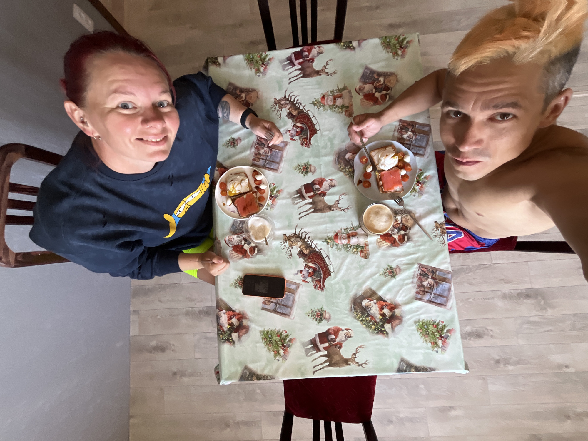 A high-angle shot shows two people sitting at a table covered with a Christmas-themed tablecloth. The person on the left is wearing a navy long-sleeved shirt and has red hair; the person on the right is shirtless and has dyed blonde hair. Both are eating a meal consisting of bread, tomatoes, and white cheese, with coffee nearby. A black smartphone lies on the table. The location is Chaikovsky, Russia.
