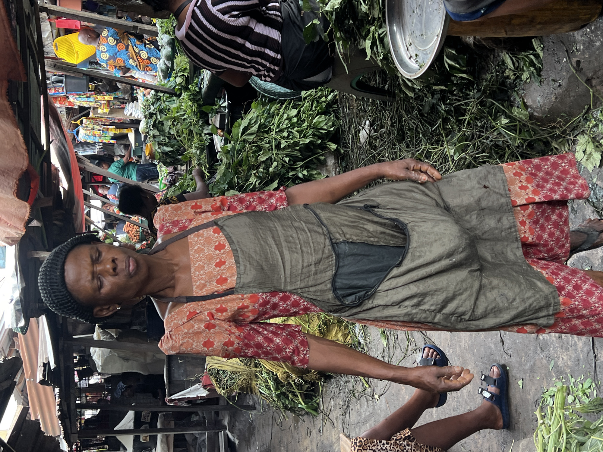 In Nkpor, Nigeria, a woman stands in a bustling market, her gaze directed downwards with her eyes closed. She wears a dark knitted cap, an orange and red patterned dress, and a grey apron. To her left, the legs of another person are visible, wearing blue flip-flops and a skirt with a leopard print. The background reveals stalls with various goods, including large piles of green vegetables, sacks, and colorful items hanging from structures. The ground is paved and appears damp. Other individuals are visible in the background, engaged in market activities.