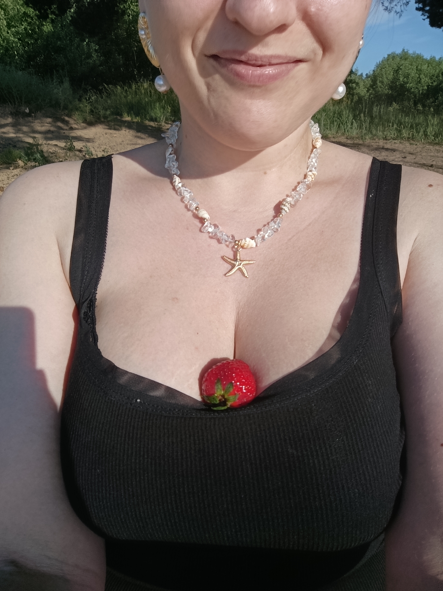A medium shot shows a person wearing a black ribbed tank top with a sheer neckline. A single strawberry rests nestled in the cleavage of the tank top. The person is also wearing a seashell and clear bead necklace with a small gold starfish charm. Large pearl earrings dangle from their ears. The background is blurred and appears to be outdoors, with green trees and foliage visible, along with a sandy or dirt path. The lighting suggests it is daytime, likely sunny. The person's face is partially visible, with a smile and their lips slightly parted. There is no visible text in the image.
