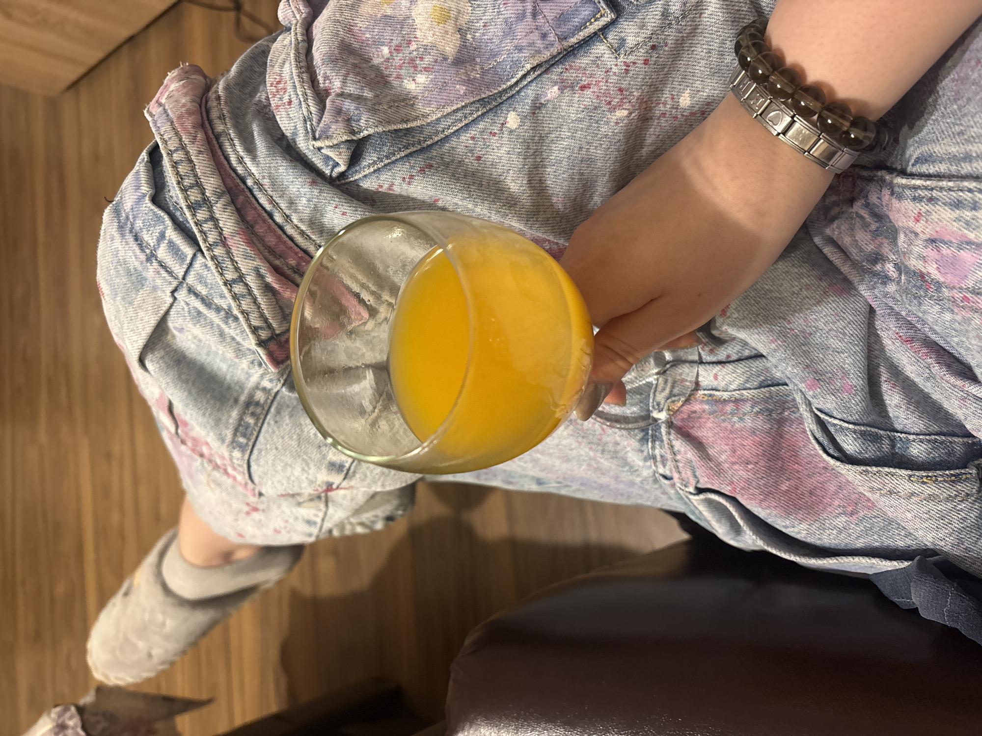 A person is holding a glass of orange juice. The person is wearing a light blue denim jacket with purple splotches, a bracelet of gray and silver beads on their right wrist, and a gray slipper on their left foot. They are seated on a dark brown chair. The background is a wooden floor and a wall. The image does not provide any information about the location being in Varna, Bulgaria.