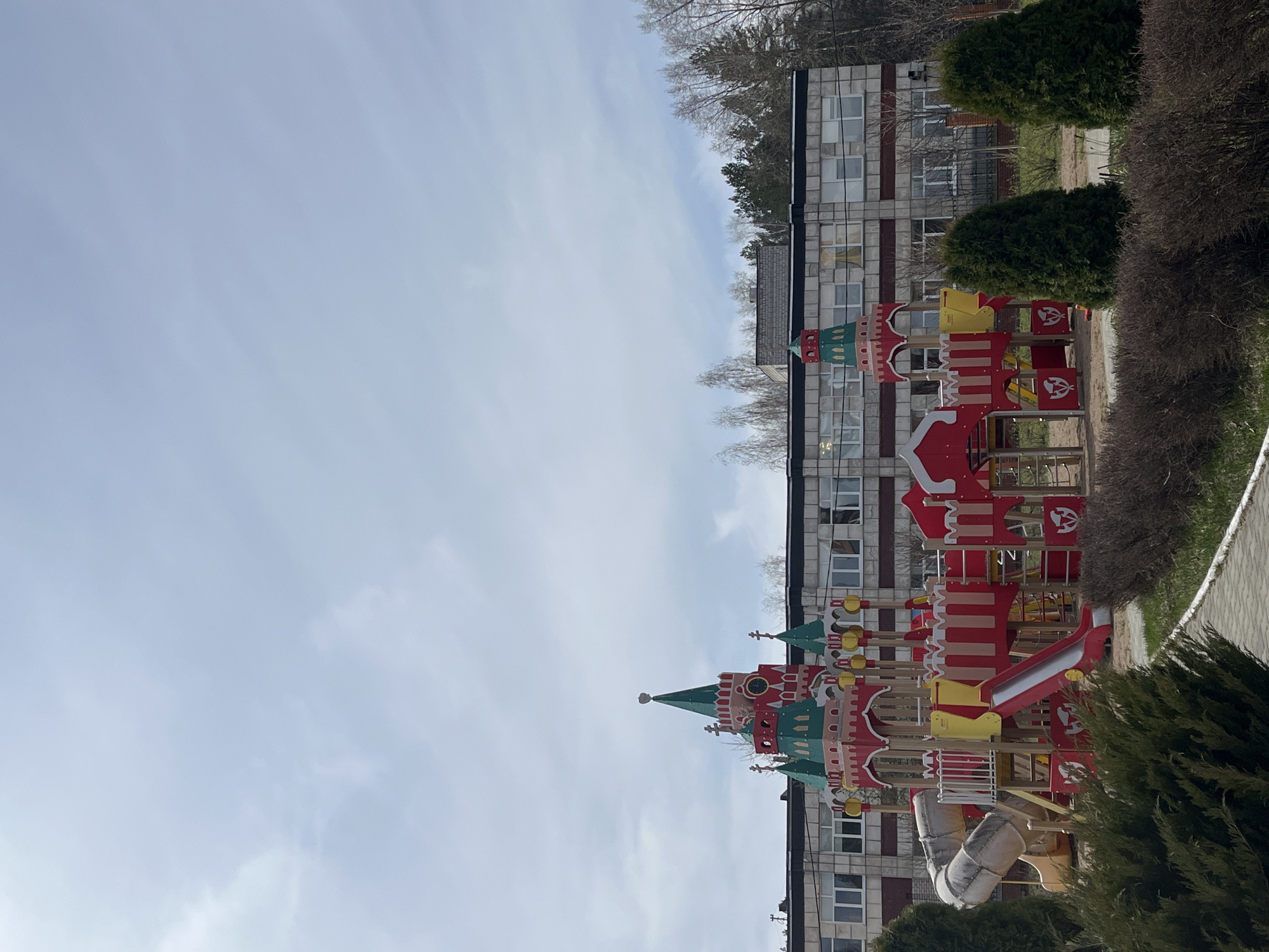 The image captures a daytime scene of a vibrant children's playground set against a backdrop of a residential building and natural foliage.
The main subject is a large, elaborate playground structure, resembling a castle, located on the right side of the frame. It features multiple sections in bright red, yellow, and green, including towers with pointed, often green, roofs and decorative elements. Visible components include an open red slide and an enclosed beige tube slide, as well as various climbing areas and platforms. Small white symbols or logos, possibly depicting stylized heads or masks, are discernible on some of the red panels of the playground structure, but no clear, readable text is present. The playground appears empty, with no children or adults in view.
Behind the playground, a multi-story building, likely an apartment complex or institutional building, stands. It has a facade of light-colored bricks, possibly concrete, interspersed with horizontal dark bands, and features numerous rectangular windows. Further back, above the building, trees with bare branches are visible, alongside some evergreen trees, suggesting a season of late winter or early spring. Green bushes and shrubs are present in the foreground and along the right side of the playground area, with a paved path or sidewalk running alongside them.
The sky occupies the upper left portion of the image and is a light blue with scattered wispy white clouds, indicating clear to partly cloudy weather and daytime lighting. The scene depicts a quiet, modern urban or suburban setting. No specific activity or event is taking place, and the overall impression is one of stillness, characteristic of a location in Chaikovsky, Russia.