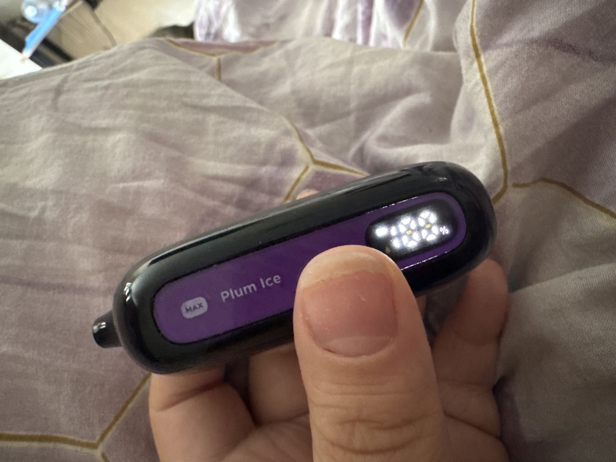 A person's thumb and index finger are visible on the left side of the image, holding a black and purple electronic device. The device has "MAX" and "Plum Ice" printed on its purple surface. A digital display on the device shows a percentage of battery life. The background consists of wrinkled, light purple fabric with a geometric pattern. The image is not in Saratov, Russia.