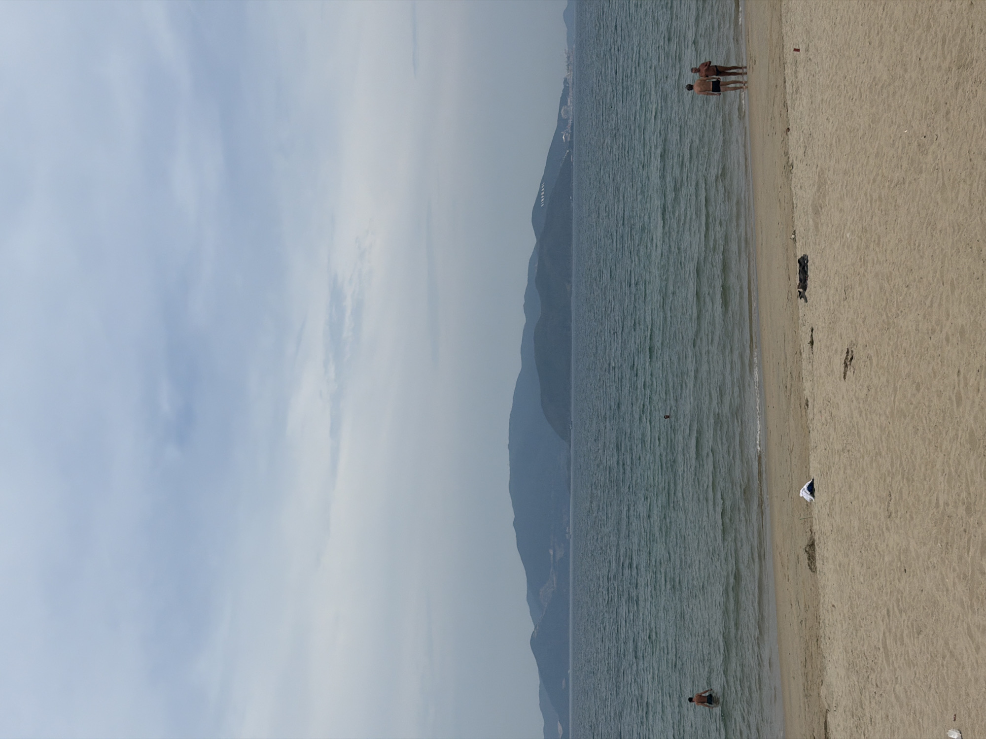 The image depicts a beach scene, likely in Nha Trang Ward, Vietnam. To the left is a view of the sky with scattered clouds. In the center is a mountain ridge along the coast. The right side shows a sandy beach meeting the ocean. There are several people on the beach and in the water. One person is walking in the water. Two other people are standing at the edge of the beach talking. There is also debris on the beach, including a white plastic bag.