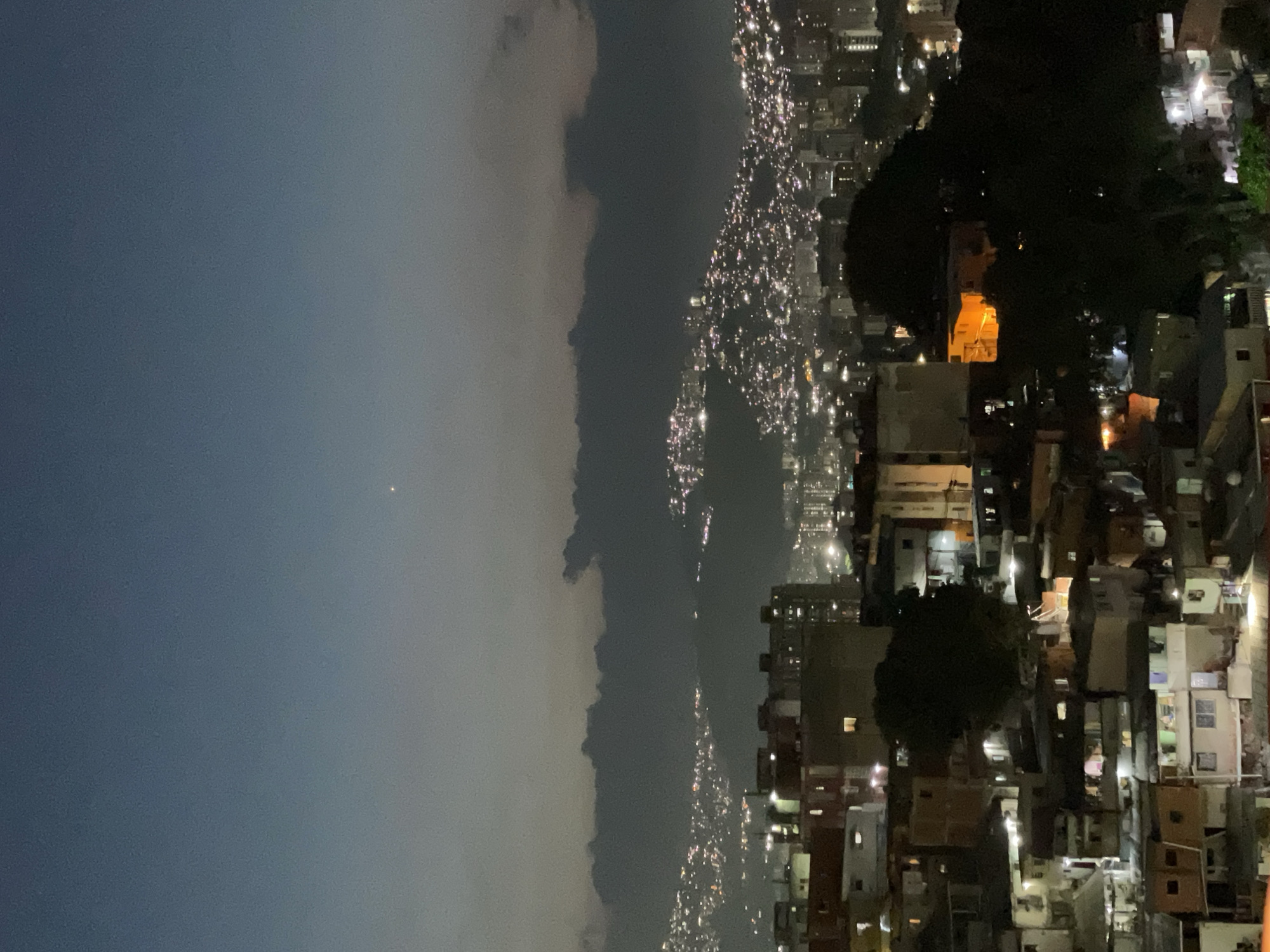The image depicts a nighttime cityscape in Caracas, Venezuela, presenting a high-angle view of a sprawling urban environment under a dark blue-grey sky. A single bright celestial object is visible in the upper central sky. A prominent band of dark, irregular clouds extends horizontally across the mid-distance, partially obscuring the upper reaches of the distant hills.
In the foreground and midground, a dense aggregation of low-rise buildings, primarily residential, occupies the lower half of the frame. These structures exhibit varied architectural styles and states of repair, with numerous windows illuminated by warm, yellow-orange light, indicating habitation. Several dark trees are interspersed among the buildings, one particularly dense cluster is centrally located on the right. A multi-story building on the mid-left shows a single illuminated window. Another building in the mid-right exhibits a distinct bright orange glow from an interior space.
Beyond these immediate structures, the terrain rises into distant hillsides heavily dotted with countless small, bright lights, indicative of extensive urban development or informal settlements (barrios) climbing the slopes. These lights extend far into the background, creating a shimmering pattern against the dark mountainous landscape. No discernible people, specific actions, or direct interactions are visible within the frame.