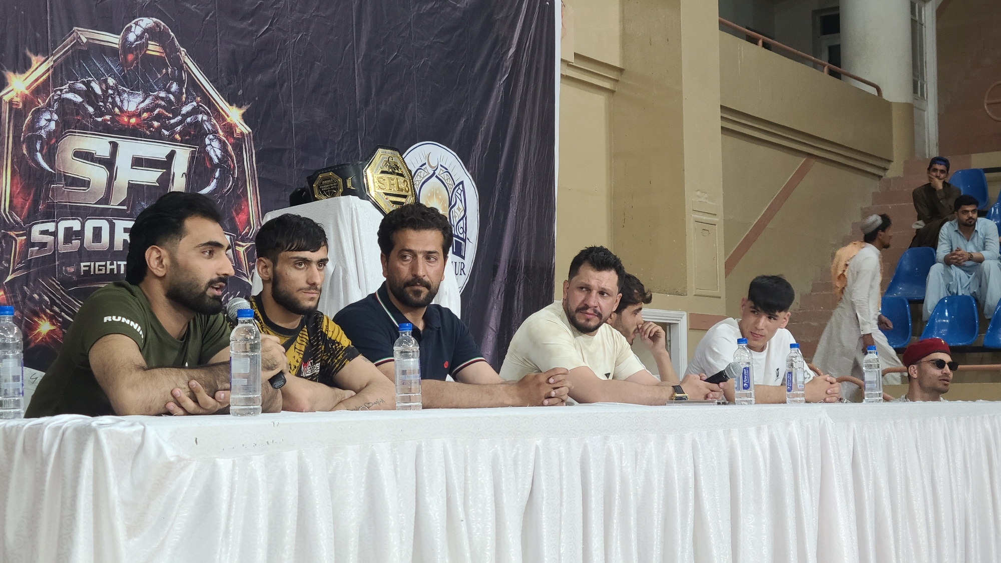 A gathering of six individuals sits behind a white tablecloth-covered table, bottles of water positioned in front of them. A banner behind them displays the logo "SFI SCORPION FIGHT." To the right, stadium-style seating is partially visible with several seated men. The location is likely Quetta City Tehsil, Pakistan.