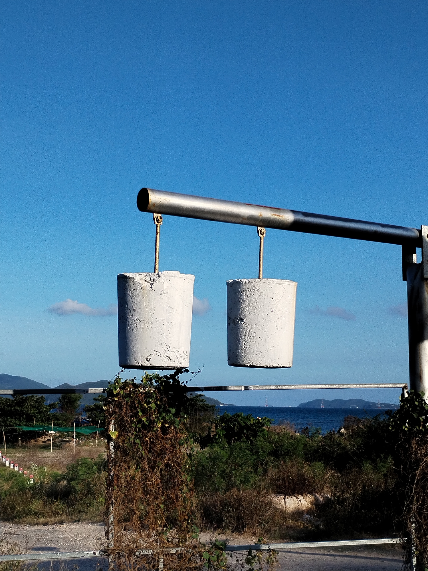 The image depicts two concrete cylinders suspended from a horizontal metal bar, likely part of a public art installation, in the Bac Nha Trang Ward, Vietnam. The cylinders, gray in color, hang from the bar by metal rods. The bar is supported by a vertical pole on the right. Below the suspended cylinders, there is overgrown vegetation, including vines clinging to a metal railing. In the background, there is a view of the sea, islands, and what appears to be a transmission tower. The sky is clear and blue.