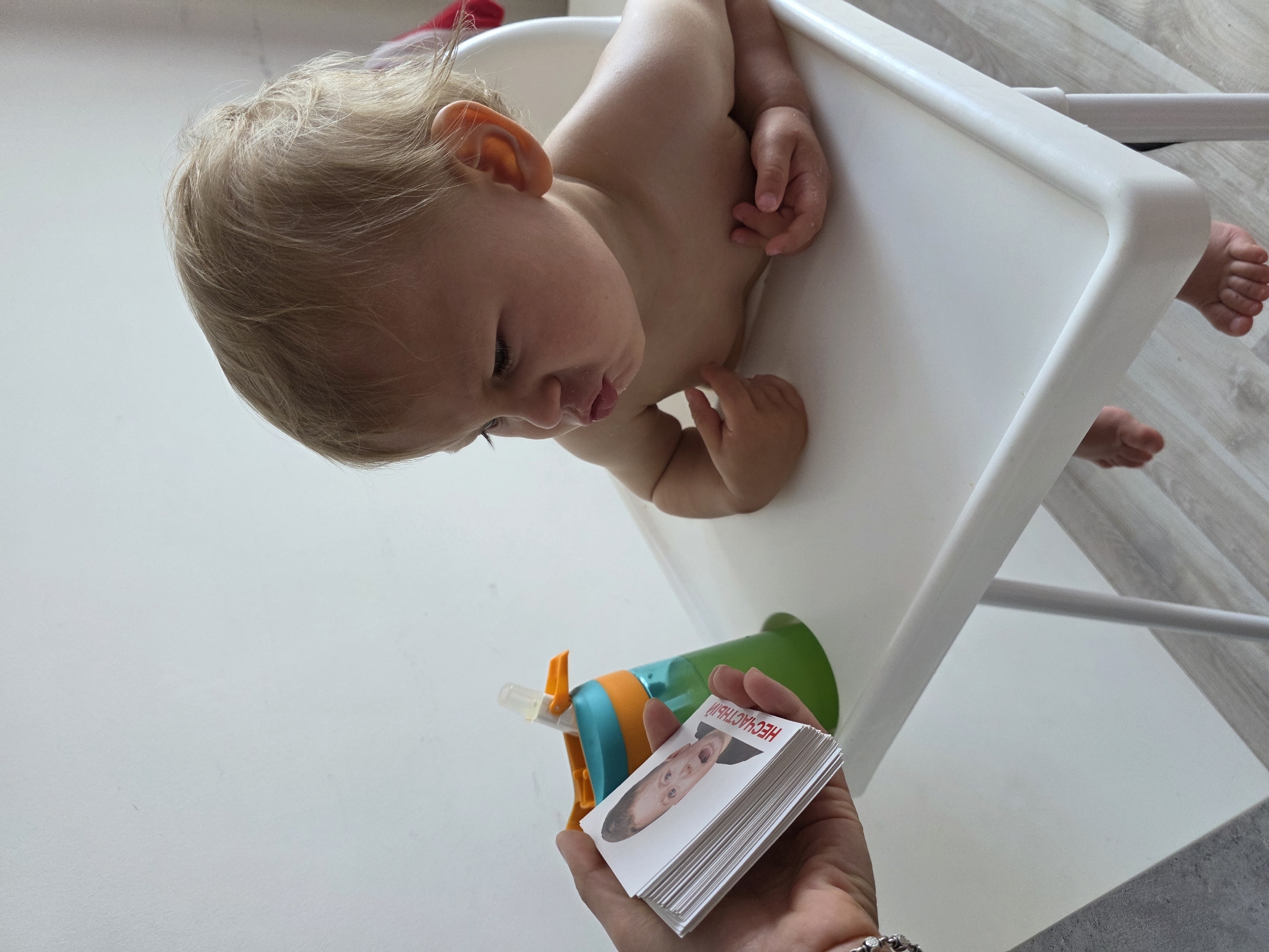 In Sochi, Russia, a baby sits in a high chair, looking at a hand holding a stack of flashcards with images of facial expressions. The baby, with blonde hair and wide eyes, pouts. A green and orange sippy cup is next to the baby. The background is a plain white wall.