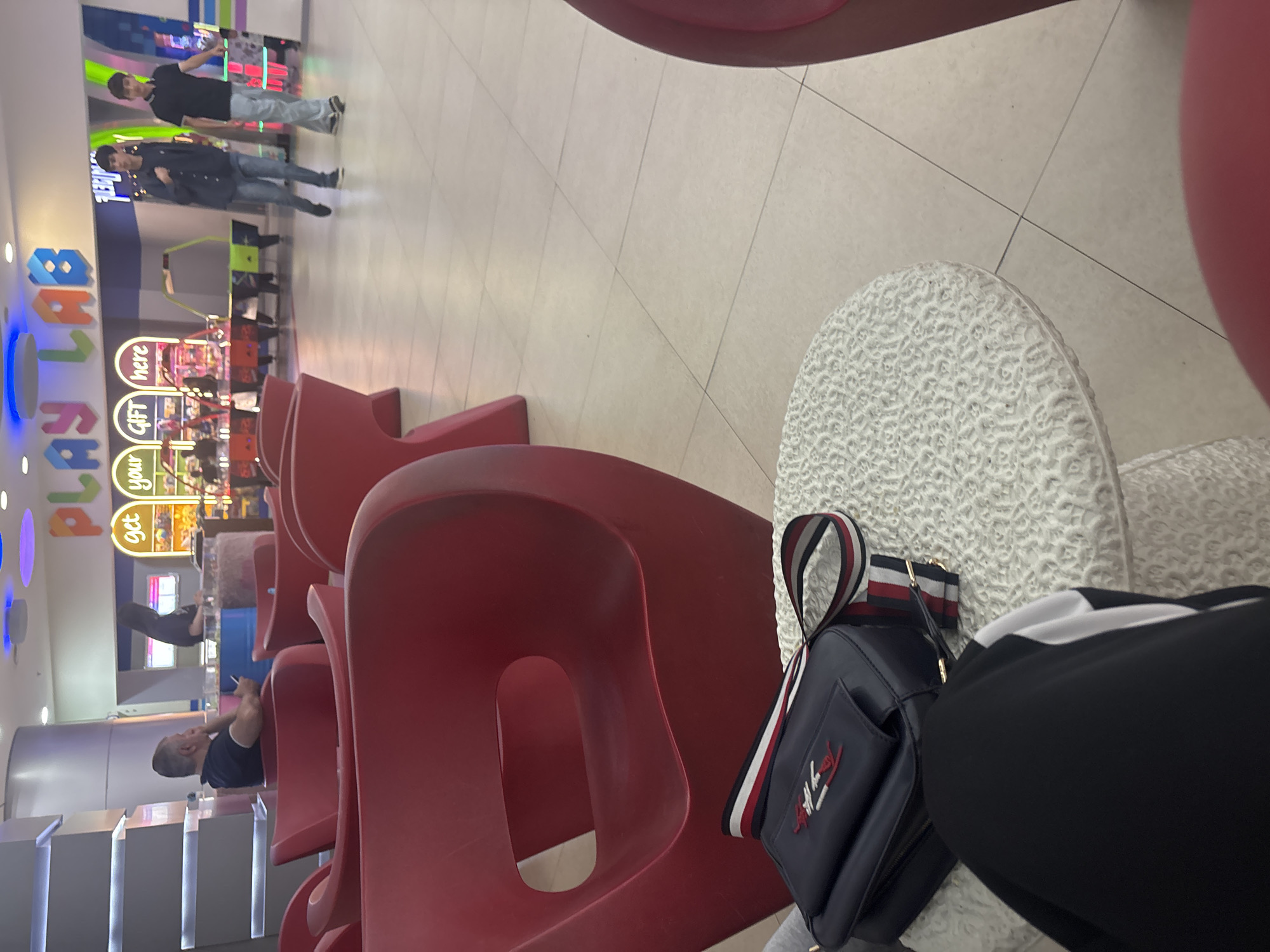 The image depicts an indoor scene within a commercial establishment, likely a mall or entertainment center, located in Yerevan, Armenia. The foreground is dominated by a waiting area featuring multiple vibrant red, molded plastic chairs, some of which are empty. A person's leg, dressed in black pants with a white stripe, is visible in the bottom right, next to a dark blue Tommy Hilfiger shoulder bag with a distinct red, white, and blue striped strap. The bag rests on a white, textured surface, possibly a small table or a patterned chair.
The background reveals a brightly lit children's play area or arcade. A large, colorful sign prominently displays "PLAYLAB," while a smaller yellow and green sign underneath reads, "get your GIFT here." Several individuals are present in this area. Two men are standing near the "PLAYLAB" sign; one wears a black t-shirt and light-wash jeans, and the other a dark jacket and jeans. Further down the seating area, another man is visible, seated in a red chair and seemingly looking at his phone. The floor throughout the space is covered in light-colored square tiles. The setting suggests a casual leisure environment.