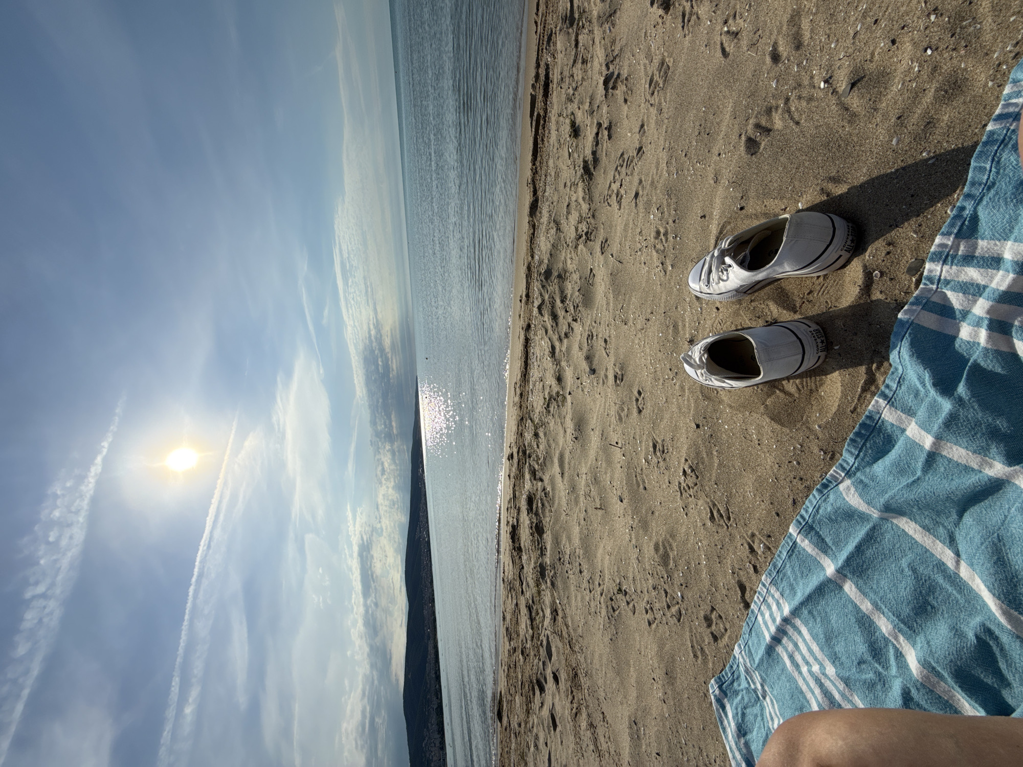 The image depicts a serene beach scene in Nesebar, Bulgaria. The foreground is dominated by a blue and white striped beach towel, partially covering a person's legs. Beside the towel, on the sandy beach, are a pair of white Converse sneakers. The sand is textured with footprints and small shells. In the mid-ground, the calm sea stretches towards the horizon, reflecting the bright sun. The sun is high in the sky, casting a brilliant light and creating a shimmering effect on the water. A distant coastline with hills and buildings, characteristic of Nesebar, is visible under a partly cloudy sky. The overall impression is one of relaxation and enjoyment of a sunny day at the beach.