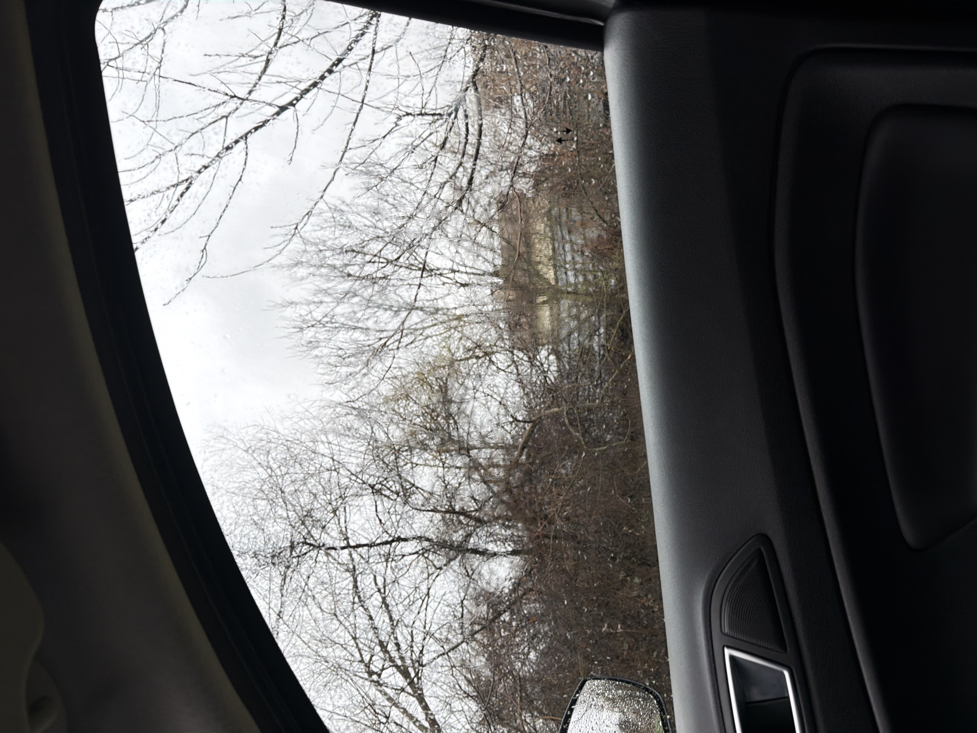 The image shows a view from inside a vehicle, likely a car, looking out of a window at a scene in what appears to be the winter season. Outside the window, there are bare trees without leaves and what appears to be either light rain or moisture on the glass. A building structure is partially visible through the trees. The interior of the vehicle is dark, with the window frame and a portion of the car's interior door panel visible. There are no people visible in the frame. It is not possible to verify if the location is in Dragichevo, Bulgaria from the image.