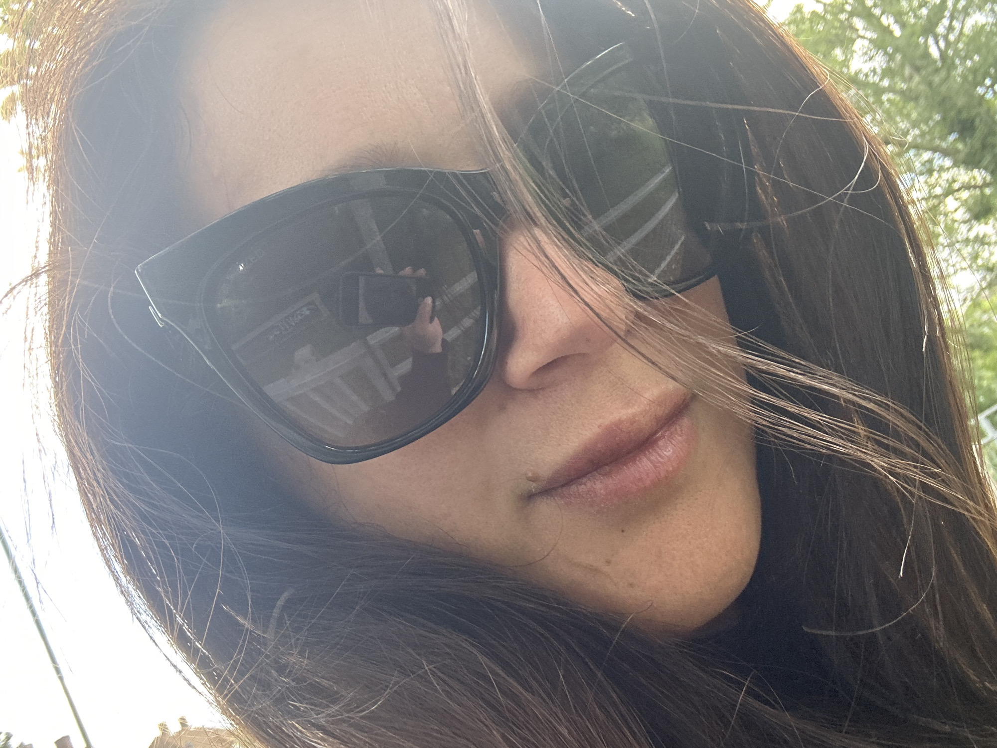 This is a close-up selfie of a woman's face, taken from a low angle. She is wearing large, dark, square-shaped sunglasses that cover her eyes. Her hair is long, dark brown, and slightly windswept. The sunlight is bright, casting a glare on the sunglasses and creating lens flare in the image, suggesting it is daytime.
In the reflection of her sunglasses, a mobile phone is visible in her hand, indicating she is taking the selfie. The background in the reflection appears to be an outdoor setting with a white railing and some greenery. The woman's lips are slightly parted, and she has a mole on her chin. Her skin tone is fair. The overall mood is casual and outdoorsy. The time of day is likely sunny, given the bright light and glare. No text is visible in the image.