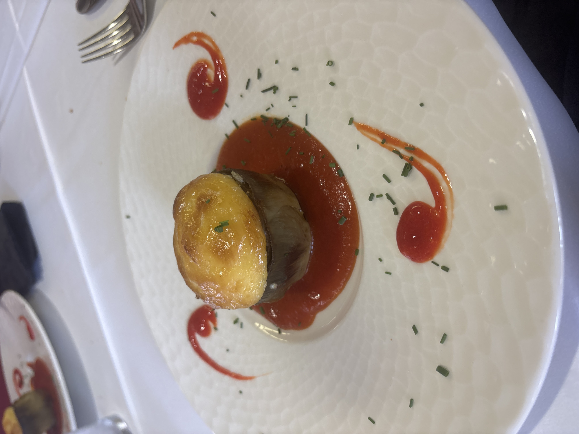 A close-up, eye-level shot captures a single serving of stuffed eggplant on a white plate. The eggplant, a cylindrical piece with a golden-brown, pastry-like topping, rests in a pool of vibrant red sauce. The plate itself features a subtle, textured pattern. Red sauce is artfully swirled onto the plate in decorative patterns, and finely chopped chives are sprinkled around the dish. The setting appears to be a restaurant in Urbanización Roquetas De Mar, Spain, suggested by the white tablecloth and the presence of a fork on the right side of the frame, indicating a dining environment. The lighting is bright, focusing attention on the food presentation.