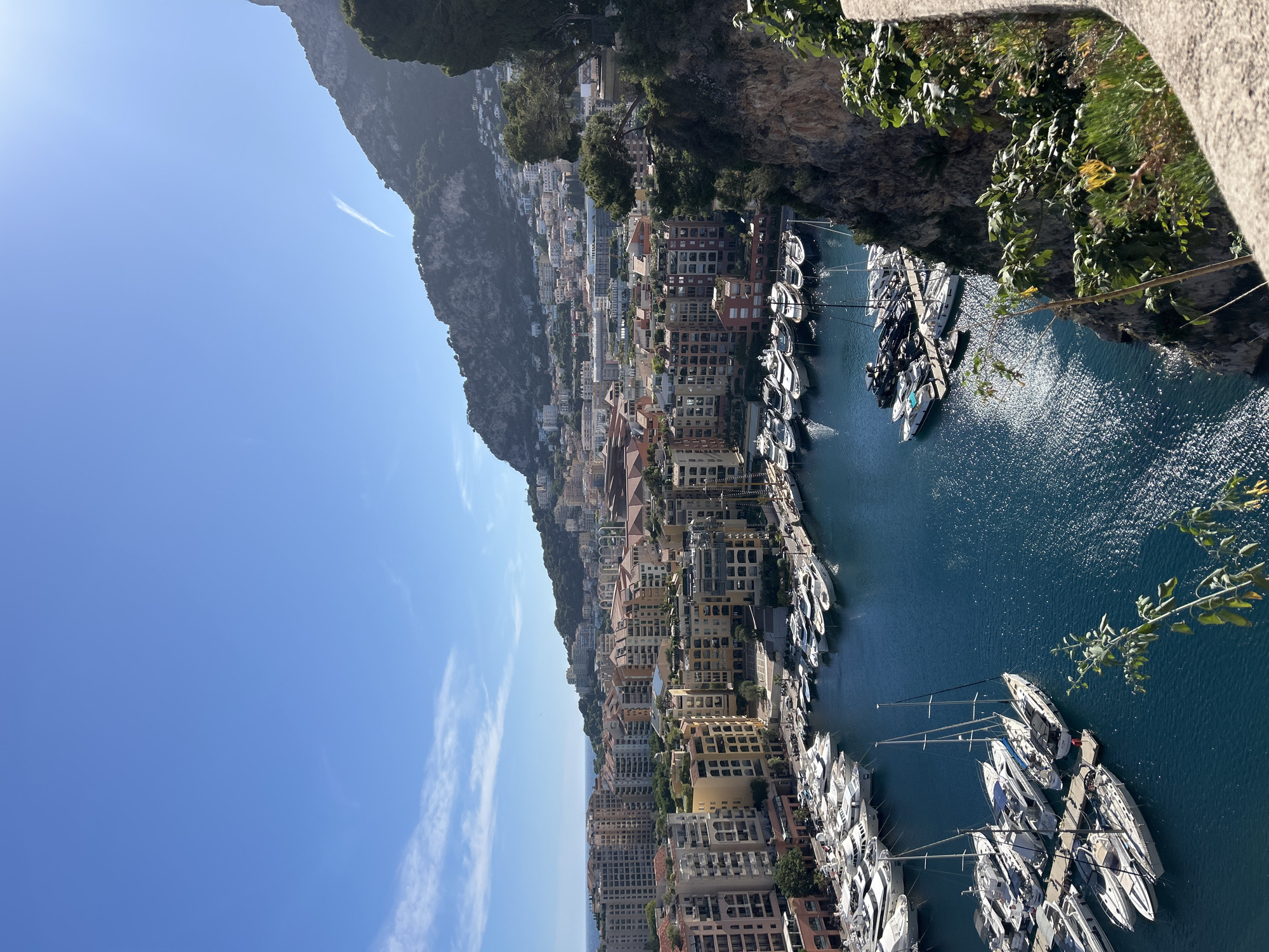 This is a high-angle, full shot of a harbor in Monaco, Monaco. The harbor is filled with numerous white yachts of various sizes docked in the deep blue water. Behind the harbor, there is a densely populated city with buildings of varying heights and architectural styles, predominantly in shades of yellow, beige, and terracotta. The city is built on the side of a steep, green-covered mountain that dominates the background. The sky is a clear, bright blue with a few wispy white clouds and a faint airplane contrail. The image conveys a sense of opulence and a Mediterranean coastal atmosphere.