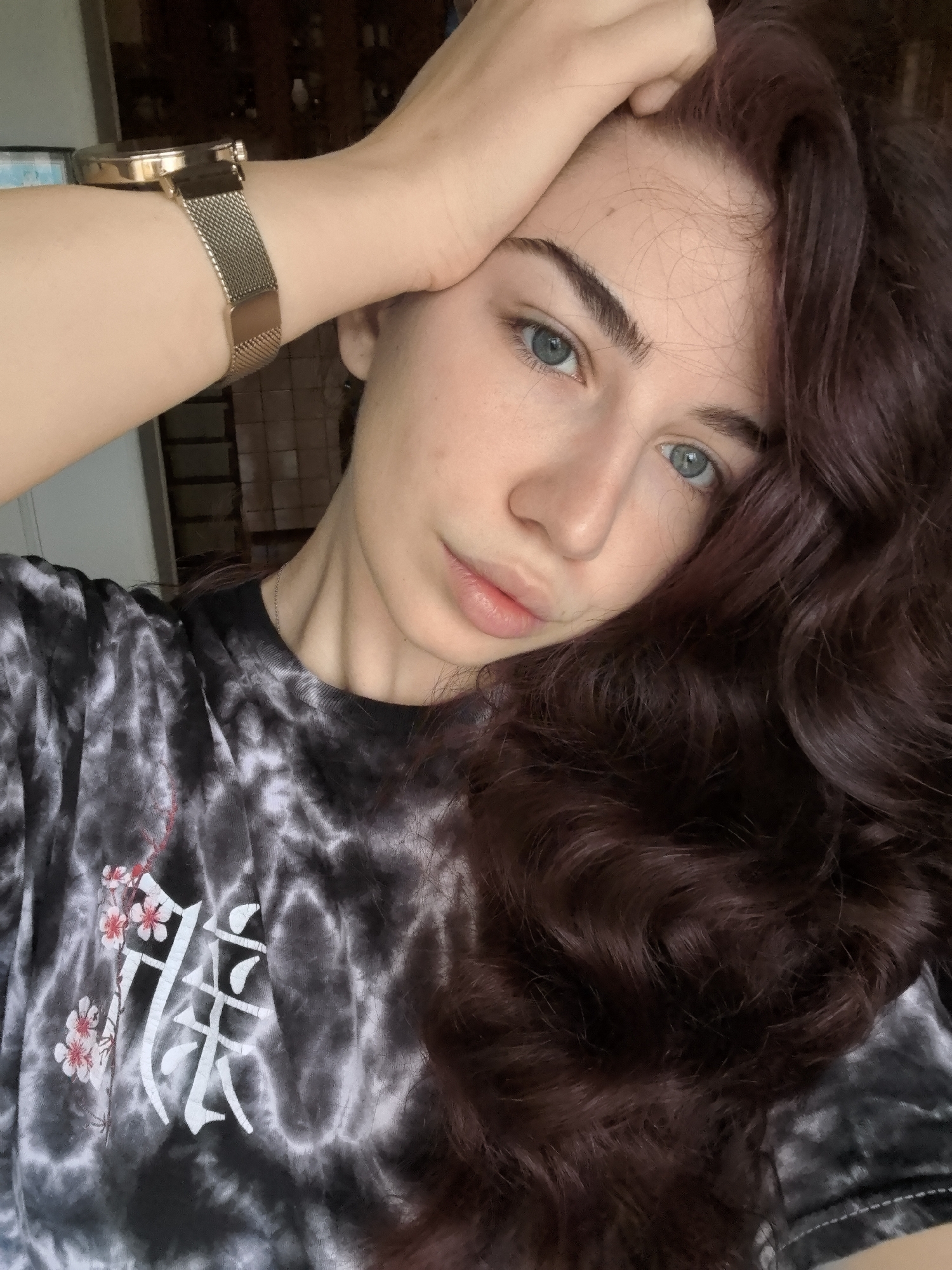 The scene depicts a young woman posing indoors in what is identified as Stara Zagora, Bulgaria. The subject is a light-skinned female with blue eyes, dark eyebrows, and light pink lips, looking directly into the camera. Her long, wavy, dark reddish-brown hair is parted on the left, flowing down to obscure the left side of her chest. Her right hand is placed on top of her head, with a gold-toned watch featuring a mesh strap visible on her right wrist. She wears a black and grey tie-dye t-shirt with a white graphic design on the left chest area, incorporating stylized Japanese characters and pink cherry blossoms. A thin silver-colored chain is visible around her neck. The background is blurred, suggesting an indoor setting with a darker area behind her head, possibly a cabinet or shelves with indistinct objects, and a lighter wall segment to the left.