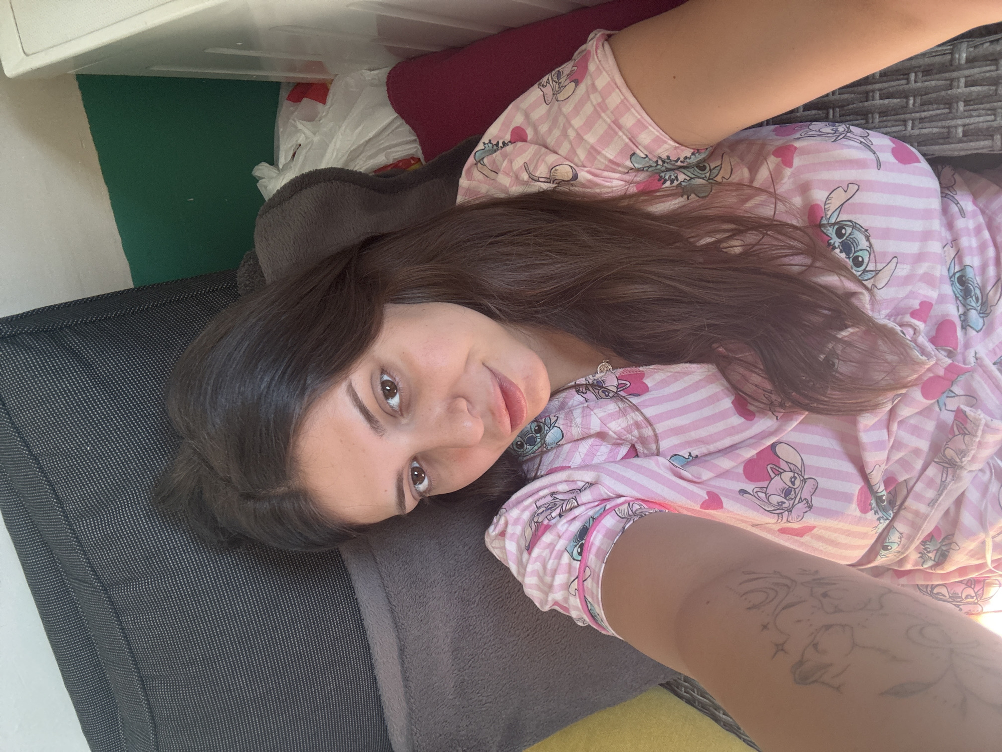 A young woman with long, dark brown hair and brown eyes is lying down, looking at the camera. She is wearing a pink and white striped pajama top with Stitch characters on it. Her left arm is visible, with a tattoo of what appears to be a deer and stars. The background consists of pillows, a green wall, and some plastic bags. This image was taken in Plovdiv, Bulgaria.