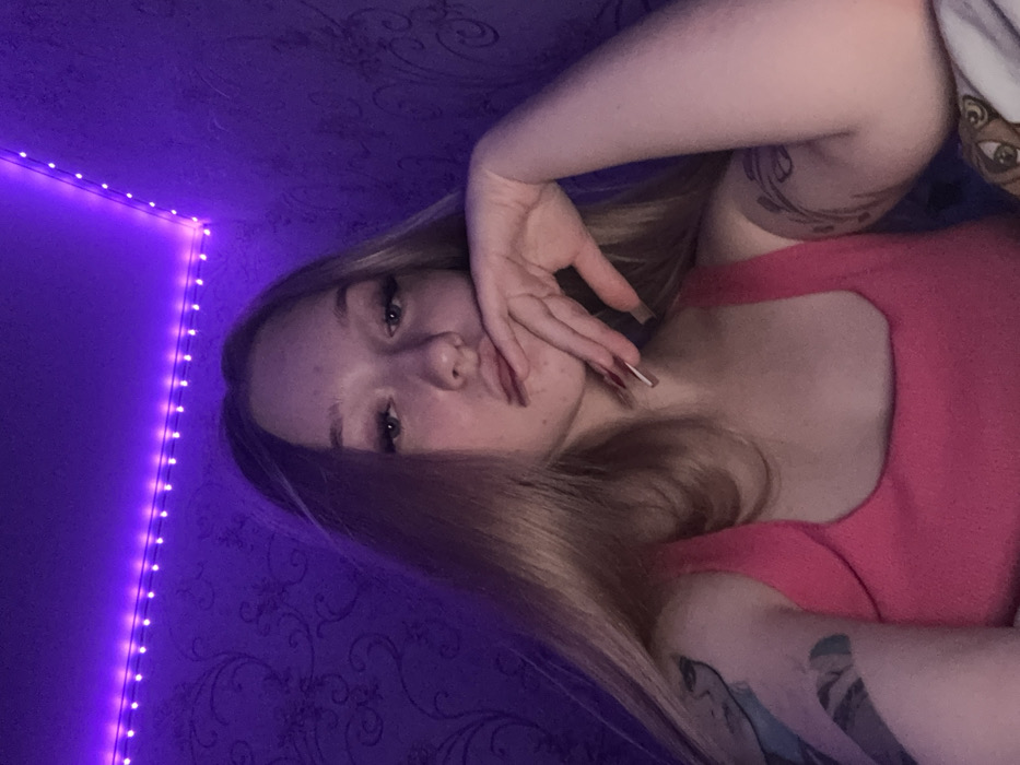 The image displays a young female subject positioned centrally in an indoor environment. She has long, straight blonde hair and is wearing a pink tank top. Her gaze is directed towards the camera. Her left hand, adorned with long, red-tipped fingernails, is raised to her face, with fingers touching her lower lip and chin. A multi-colored tattoo is visible on her left upper arm, and a dark, patterned tattoo is visible on her right upper arm. The background consists of a dark purple wall with a subtle patterned wallpaper, illuminated by a strip of purple LED lights along the ceiling-wall corner. The lighting is predominantly dim and purple. A patterned fabric is partially visible in the bottom right corner of the frame. The specific location within the city of Saratov, Russia, is not ascertainable from the visual information provided in the image.
