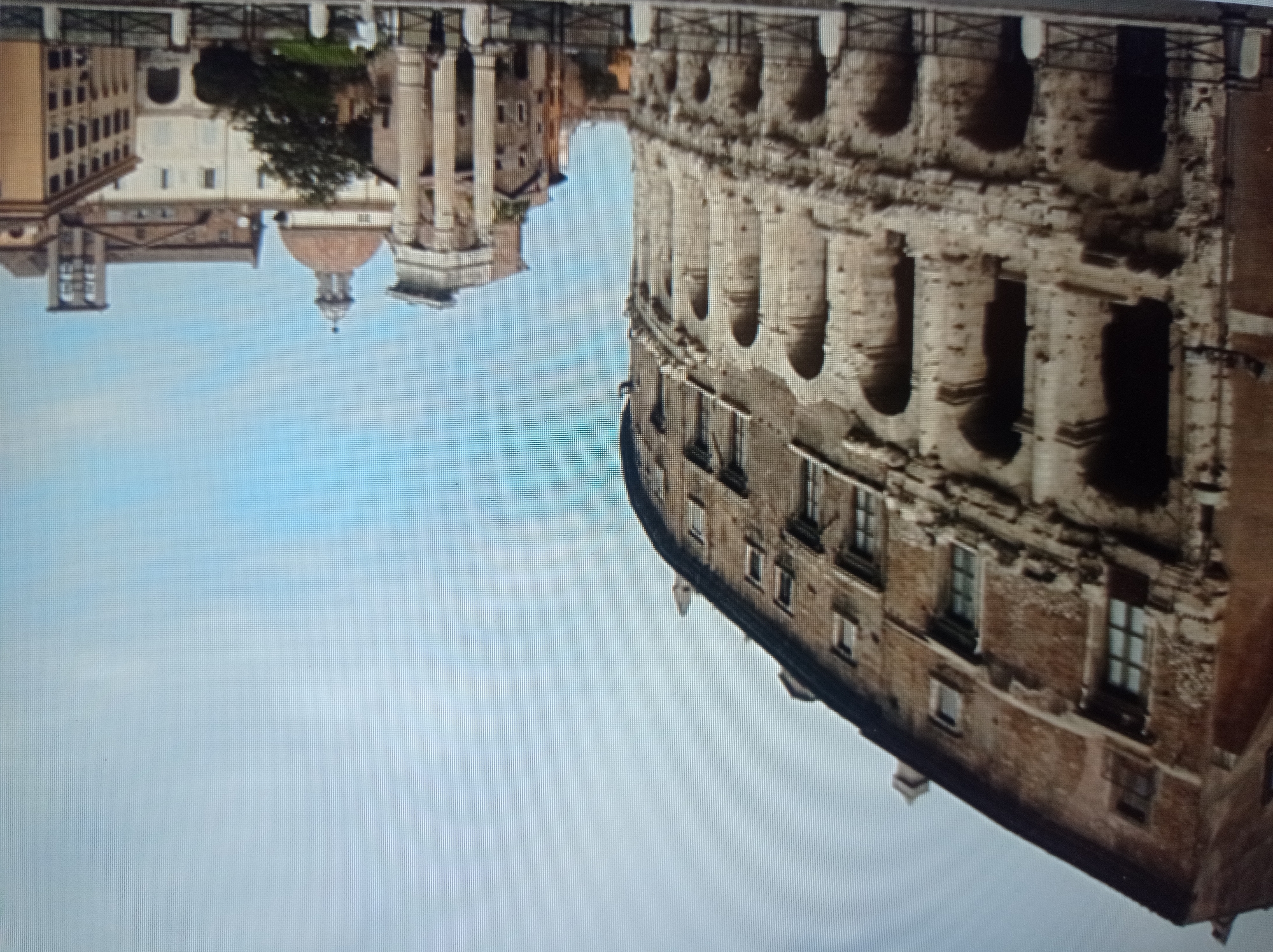 The image depicts the Theater of Marcellus in Rome, Italy. The ancient Roman amphitheater, characterized by its weathered stone arches and multi-story structure, occupies the left side of the frame. In the background, various buildings with classical architectural elements, including columns and a dome, are visible. The sky is overcast and a pale blue. A few people are present, standing in front of the theater and near the background buildings. The scene is set outdoors in what appears to be an urban environment. There is no indication that this location is in Mukachevo, Ukraine.