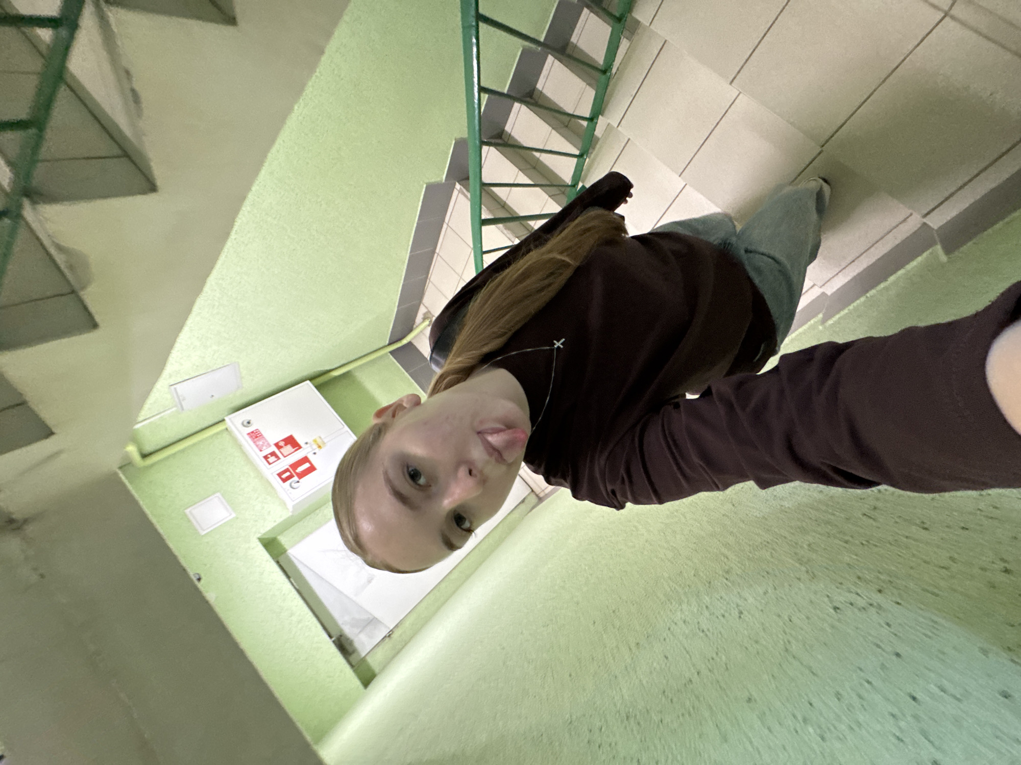 A young woman with fair skin and blonde hair is taking a selfie in a stairwell. She is wearing a dark-colored long-sleeved shirt and jeans. The stairwell has green walls and metal railings. A fire extinguisher box is visible on the wall. The stairwell is located in Perm, Russia.
