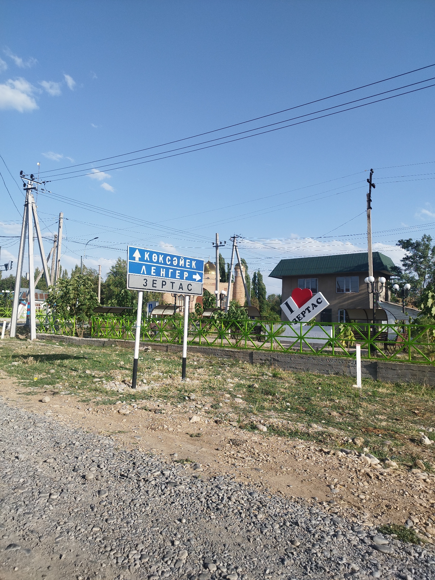 The image depicts a street scene in Зертас ауылдық округі, Kazakhstan, on a sunny day. In the foreground, a gravel road leads towards a green metal fence. Beyond the fence, a signpost displays directions in a Cyrillic script. The upper part of the sign has an upward arrow and the word "КӨКСУЕК". The middle section has a rightward arrow and the word "ЛЕНГЕР". The bottom section has the word "ЗЕРТАС". To the right of the signpost, a large 3D "I LOVE ЗЕРТАС" sign is visible, with a red heart placed above the word "LOVE". A building with a green roof is situated behind the "I LOVE ЗЕРТАС" sign. Power lines and utility poles are prominent in the background. The environment is semi-rural, with trees and vegetation present. No people are visible in the image.