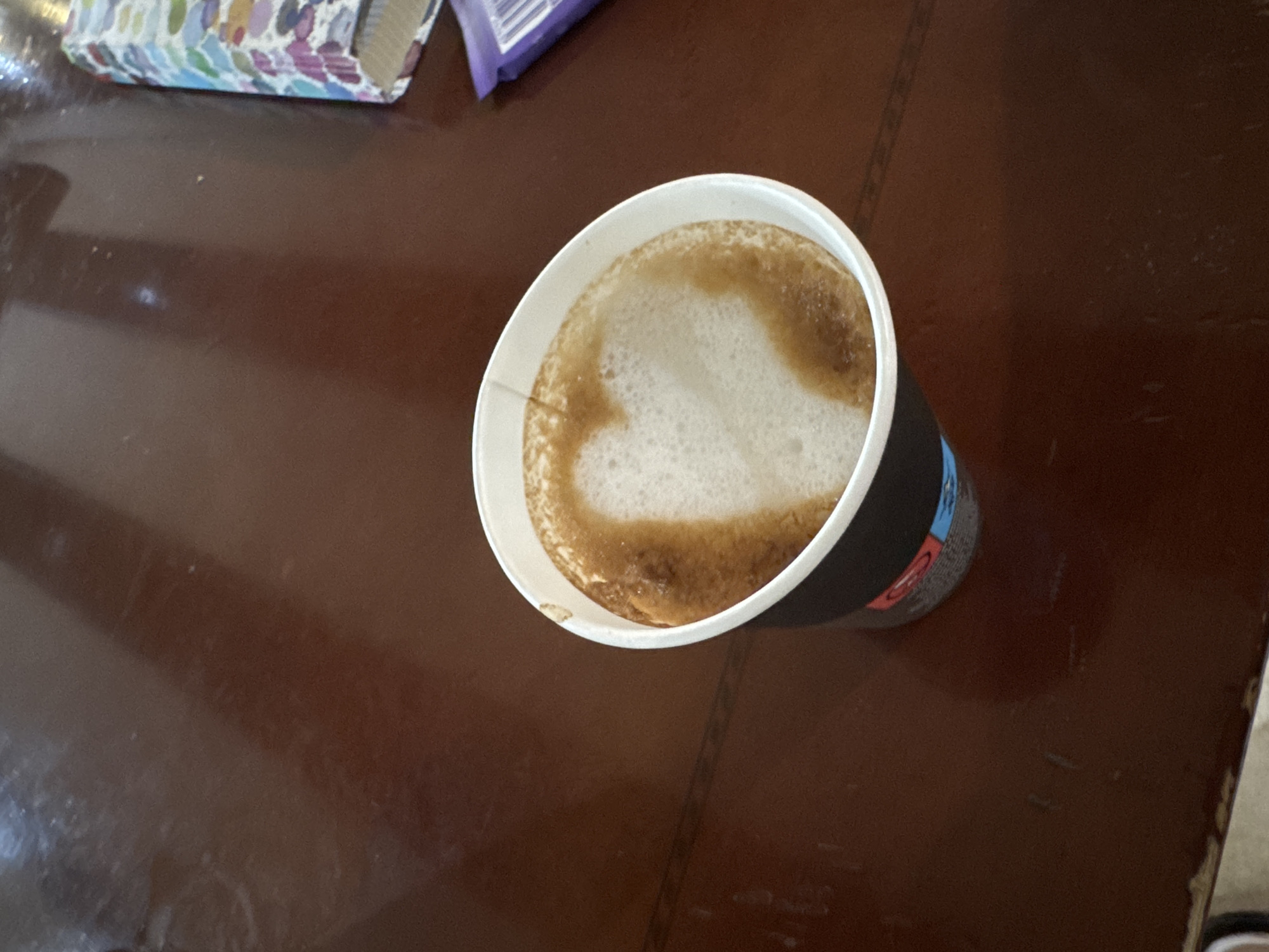 A close-up, high-angle shot shows a disposable coffee cup with a heart shape latte art on a dark brown wooden table. The cup is black with some branding and has a white rim. The coffee inside has a creamy white foam with a heart shape made from the coffee. The table surface is glossy and reflects some light. To the right of the coffee cup, a portion of a colorful patterned object and a purple wrapper are visible, possibly a book or a box of candy. The image is taken indoors, likely in a cafe or home setting in Sredets, Bulgaria. There are no people visible in the frame.