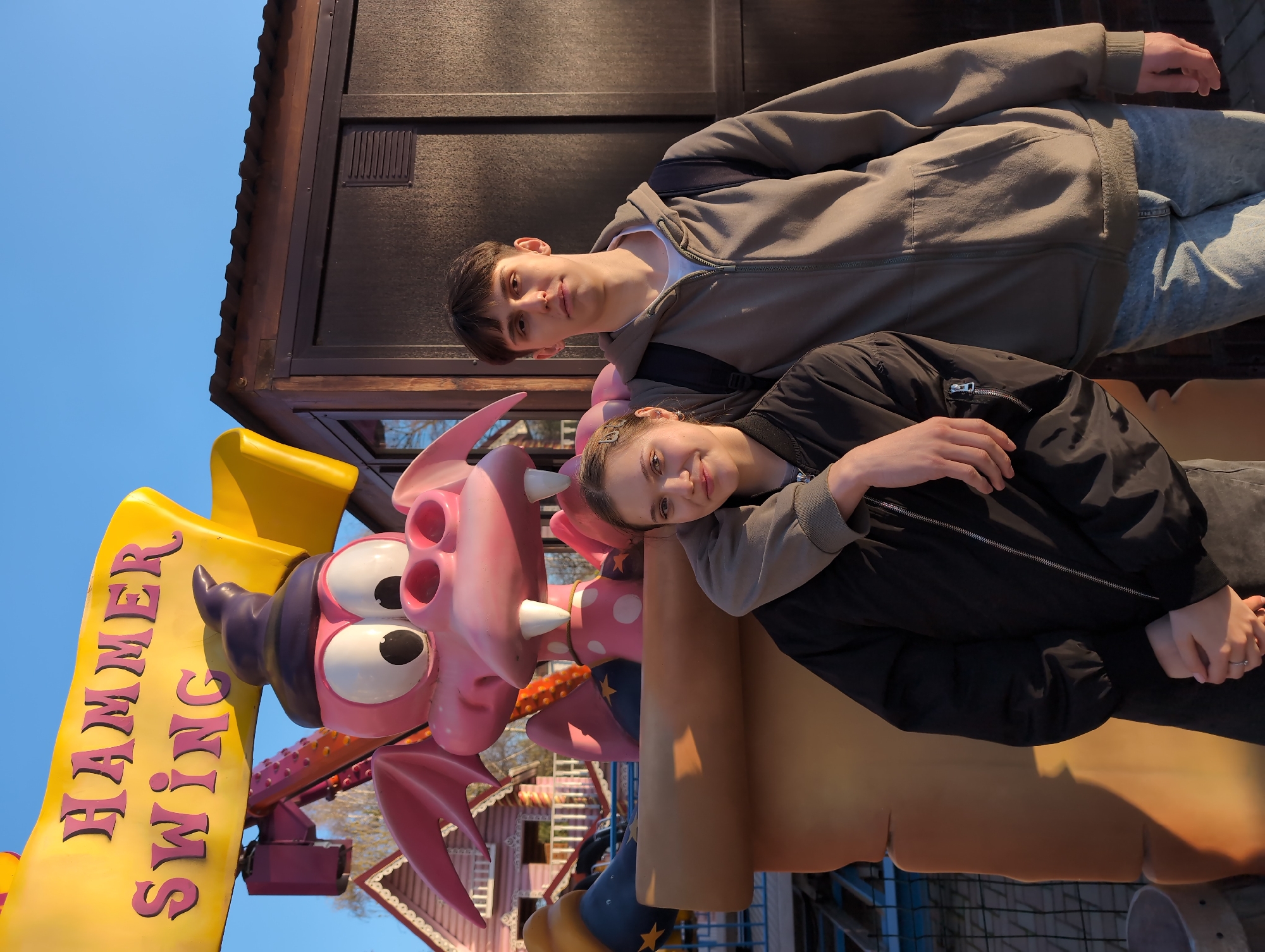 Two young people are posing in front of the "Hammer Swing" ride at the Park of Culture and Rest named after S.M. Kirov in Izhevsk, Russia. The ride features a pink dragon cartoon character with a purple hat and large yellow sign with red writing. The male is standing behind the female, and they are both looking at the camera. The male is wearing a gray zip-up hoodie, jeans, and a backpack. The female has pink streaks in her hair and is wearing a black jacket over a gray shirt. The background includes a building, fence, trees, and blue sky.