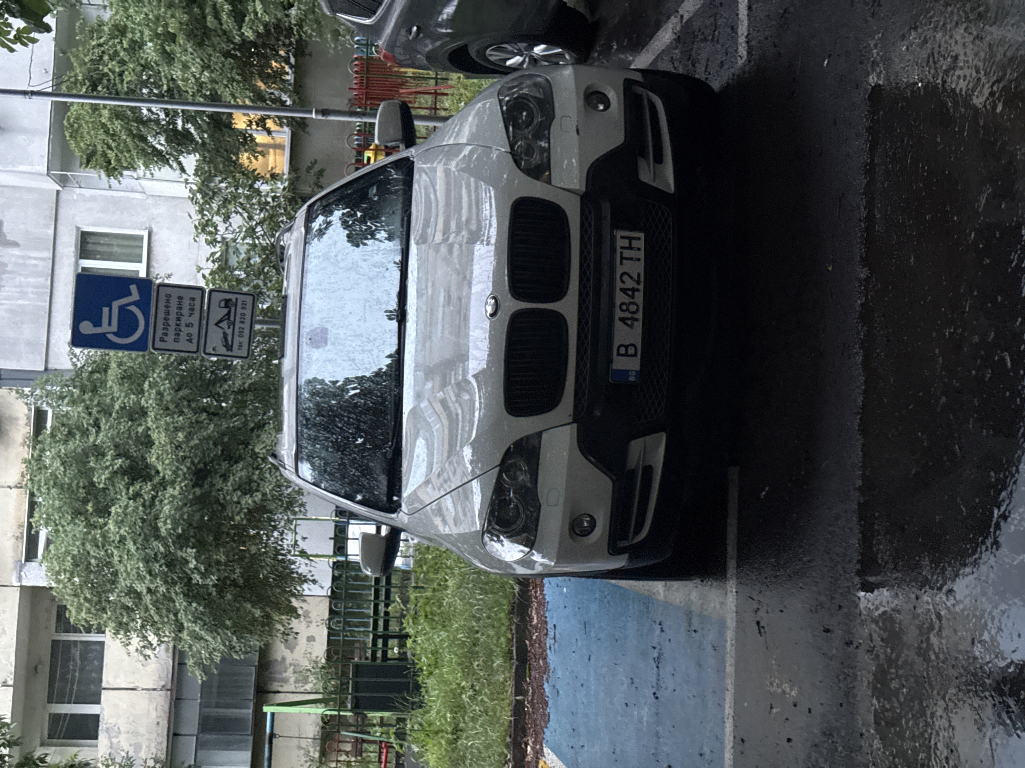 A white BMW X6 SUV is prominently featured, parked on wet asphalt in an urban setting, likely in Varna, Bulgaria. The ground's wetness and reflections indicate recent rain. The vehicle is positioned in a parking space, partially marked with blue paint, suggesting it is a designated spot.
Adjacent to the car, two informational signs are mounted on a pole. The first is a standard blue sign with a white wheelchair symbol, denoting disabled parking. Below it, a white sign with text in Bulgarian reads "Разрешено паркиране до 5 часа" (Permitted parking up to 5 hours), alongside a tow-away symbol and a contact number: "тел: 052 820 921". The "052" area code is associated with Varna, Bulgaria.
The BMW's front license plate, "BG B 4842 TH", is clearly visible, indicating it's a Bulgarian registration. While the 'B' code usually signifies Sofia, the provided context places the scene in Varna. In the background, a light-colored multi-story building with visible windows is partially obscured by lush green trees and foliage. The overall lighting is soft, indicative of an overcast day. The image itself appears slightly rotated.