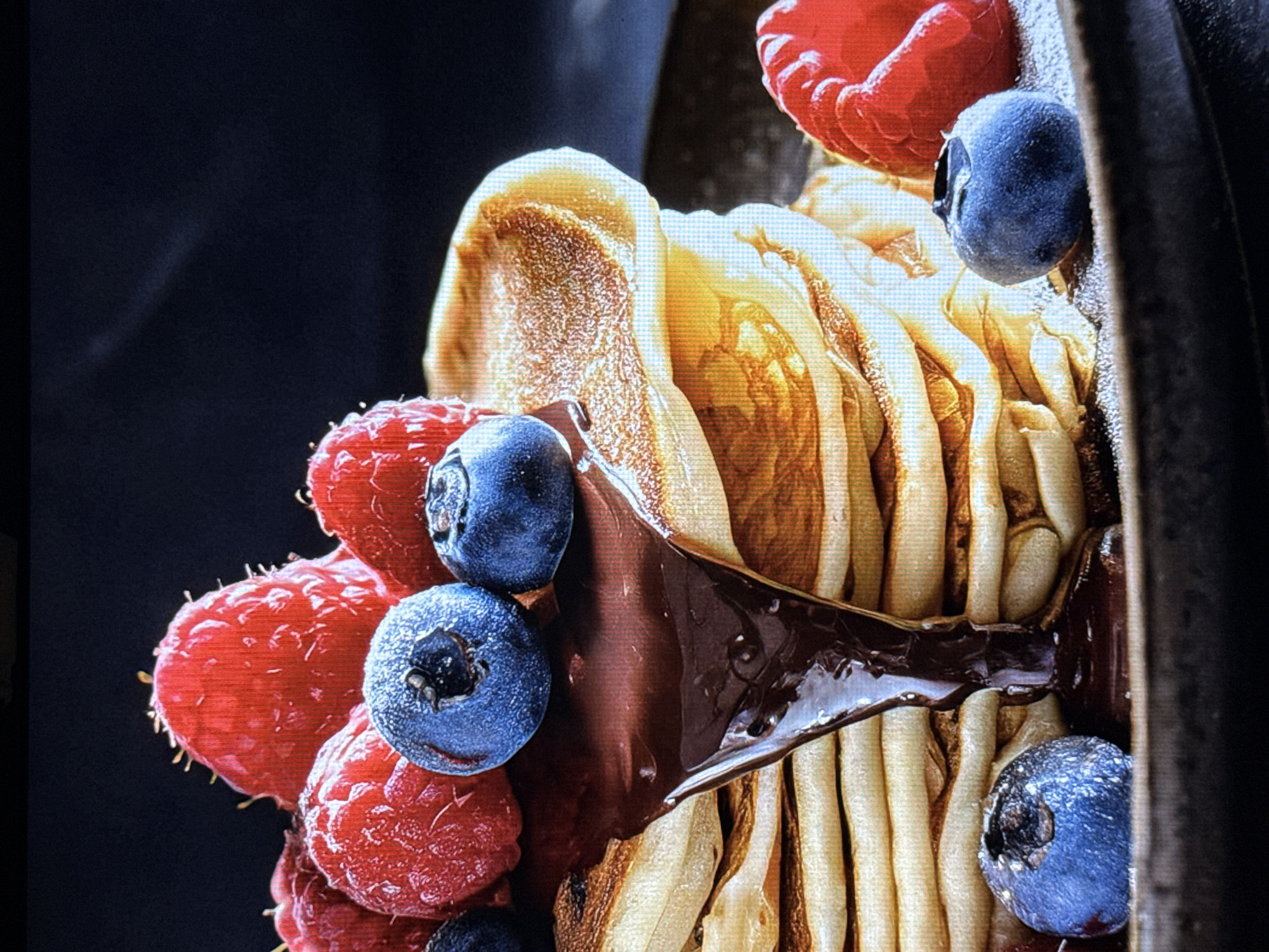 The image displays a close-up, macro shot of a dessert. The primary subjects are several ripe raspberries and blueberries, artfully arranged around a stack of what appear to be thin crepes or rolled pancakes. A dark, glossy chocolate sauce is drizzled over the crepes and fruit, adding a rich texture and visual appeal.
The raspberries are a vibrant red, with their characteristic fuzzy surface visible. The blueberries are a deep blue, with a slight powdery bloom on their skin. The crepes are a golden brown, with visible ripples and folds, suggesting a light and airy texture. The chocolate sauce is thick and reflective, catching the light.
The background is dark and out of focus, which serves to highlight the colorful and detailed subjects in the foreground. There are no people, text, or specific location cues visible in the image. The lighting appears to be artificial, designed to emphasize the textures and colors of the food. The overall impression is one of a decadent and visually appealing dessert, likely prepared for enjoyment. The focus is entirely on the food itself, presented in an inviting and appetizing manner.