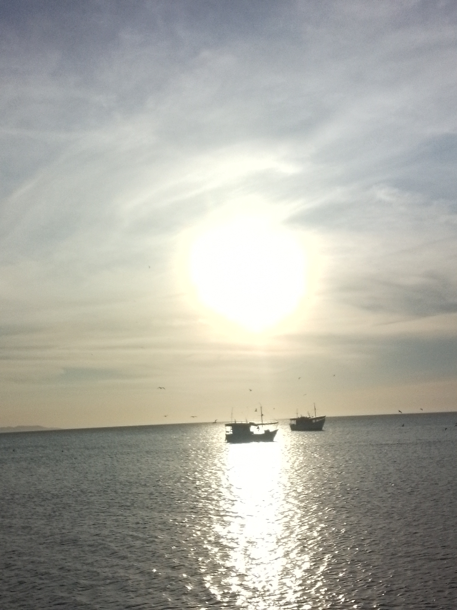 The image depicts a seascape at Juan Griego, Venezuela, under a bright, hazy sun. Two fishing boats are silhouetted on the water, their masts visible. The sun's reflection creates a glittering path on the surface of the water. Several birds are scattered across the sky. A distant coastline is barely visible on the horizon. The sky is pale with light clouds.