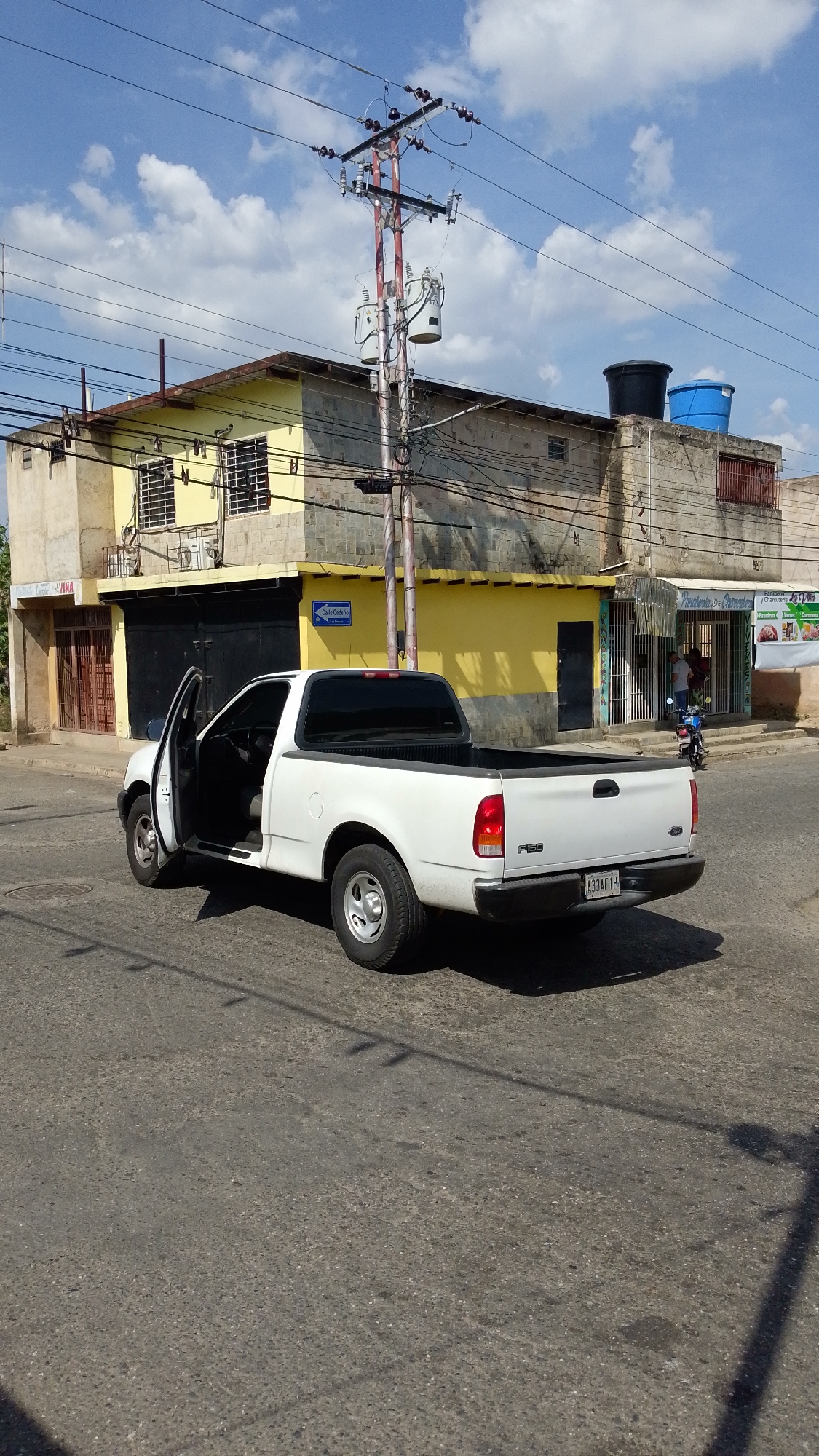 The image captures a street scene in Tinaquillo, Venezuela. A white Ford F-150 pickup truck with an open driver-side door is parked on a paved street. The license plate reads "A33AF1H". A utility pole with power lines and transformers is positioned nearby. Two buildings with a mix of yellow and concrete exteriors form the backdrop. The building on the left has a dark gate and a door to the left of the gate, while the building to the right has a water tank on the roof. A store is visible, with a person standing inside, with a scooter parked outside. A blue street sign is partially visible on the corner of the yellow building, reading "Calle Cedeno". The sky is blue with scattered clouds.