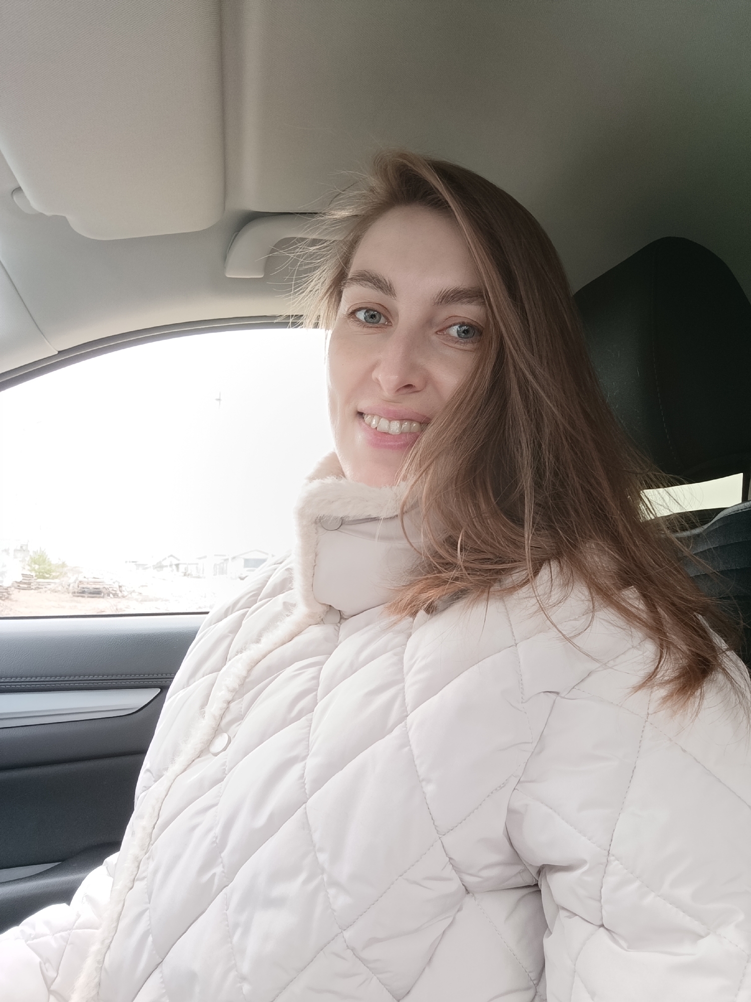 A young adult woman is captured in a close-up, chest-up selfie taken inside a car. She has long, light brown hair, blue eyes, and is smiling broadly, revealing her teeth. Her hair is slightly windswept, framing her face. She is wearing a light-colored (cream or off-white) quilted puffer jacket with a plush, possibly faux fur-lined collar, suggesting cool weather.
The setting is the interior of a vehicle, likely a modern car, indicated by the visible headliner, a sun visor, a grab handle, and a dark car seat headrest behind her. Part of a dark door panel with a silver trim element is also visible on the left.
Through the car window on the left, a bright outdoor scene is visible. The sky appears overcast or very bright, illuminating the scene. In the mid-ground, there are hints of structures or buildings, possibly in a developing area, along with what looks like stacked materials or debris. The ground seems bare, with sparse vegetation, hinting at a season like late autumn or early spring, or a mild winter without snow cover. The overall light suggests it is daytime.
The woman is looking directly at the camera with a cheerful and friendly expression. No text is visible in the image. The location is Chaikovsky, Russia.