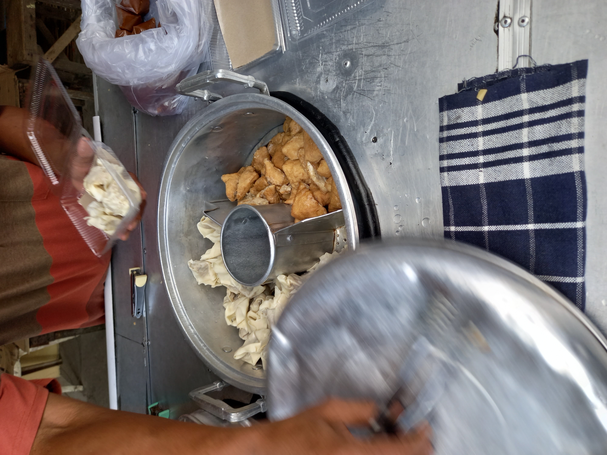 The image depicts a street food vendor in Samarinda, Indonesia. A person, partially visible and wearing a coral orange t-shirt and a brown and orange striped shirt, is preparing food on a metal cart. In the foreground, a large metal pot with compartments contains several portions of light-colored dumplings and fried brown food items, possibly tofu or tempeh. To the right of the pot, a clear plastic bag holds several small, brown, individually wrapped packages, likely a sauce or condiment. The vendor is holding a clear plastic container filled with dumplings, suggesting they are serving a customer or preparing an order. A blurred metal lid lies in the lower left of the frame, indicating recent activity. A blue and white checkered cloth rests on the cart below the pot. The overall environment suggests an outdoor, bustling food stall setting.