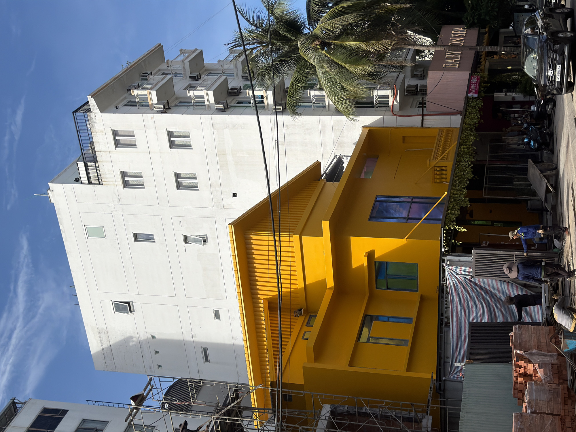 The image captures a street scene in Đà Nẵng, Vietnam. A tall white building with multiple windows and balconies stands next to a striking yellow building with a unique architectural design. A palm tree partially obscures the upper portion of the white building. The signage for "BABY ON SPA" is visible, suggesting a business establishment nearby. In the foreground, construction or renovation work appears to be underway, with construction materials like bricks stacked on the side. Several workers are present, engaged in various tasks. The sky is a clear blue, indicating daylight conditions.