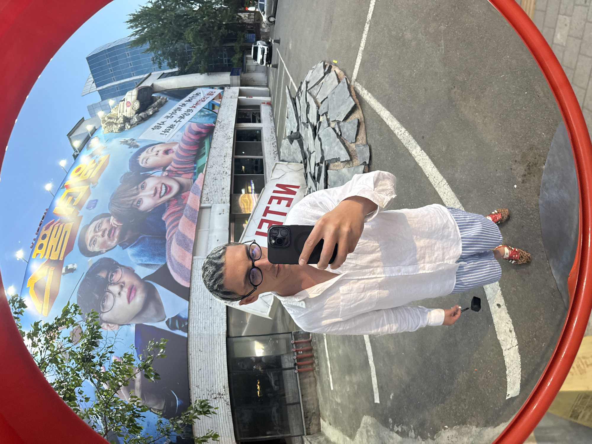 Captured in a convex mirror with a red frame, a person stands, holding a smartphone and facing the lens. They wear a white linen shirt, blue striped pants, and sandals with leopard print. Their hair is short and gray. They are surrounded by reflections of buildings, trees, and parking spaces. Dominating the backdrop is a large billboard with faces and the word "Netflix". Further back, a building appears to have a car precariously mounted on a pile of rocks. The environment suggests an urban setting. This location is in Seoul, South Korea, based on the Netflix branding in Korean.